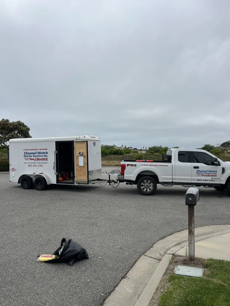 A white pickup truck towing a trailer with a Channel Watch Marine Services Inc. logo parked on the side of a road with a cloudy sky overhead. There's a trash bag on the ground near the curb.