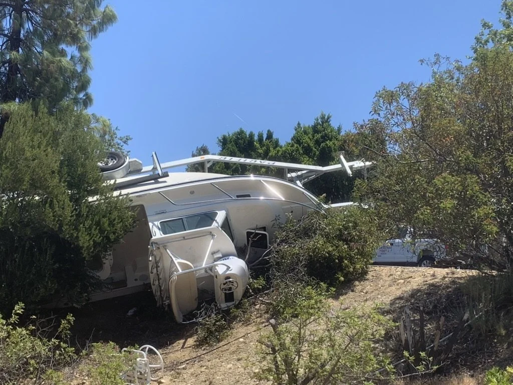 An overturned white boat on a hillside, partially hidden by bushes and trees, with a white vehicle and parts of the boat's trailer visible in the background.