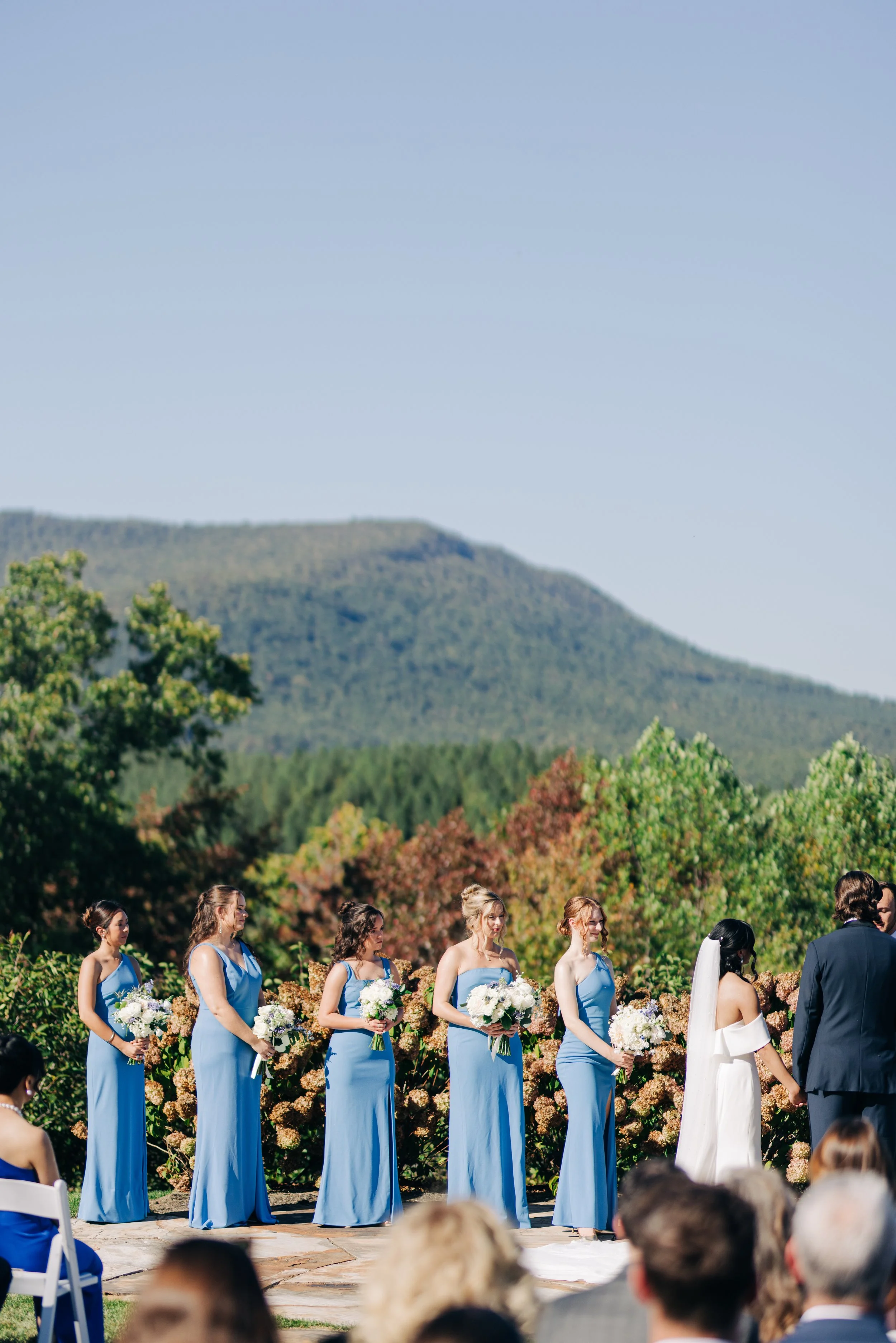 Outdoor wedding ceremony with bridesmaids in blue dresses holding bouquets, officiant, bride and groom, surrounded by guests, with a scenic mountain and forest backdrop on a clear day.