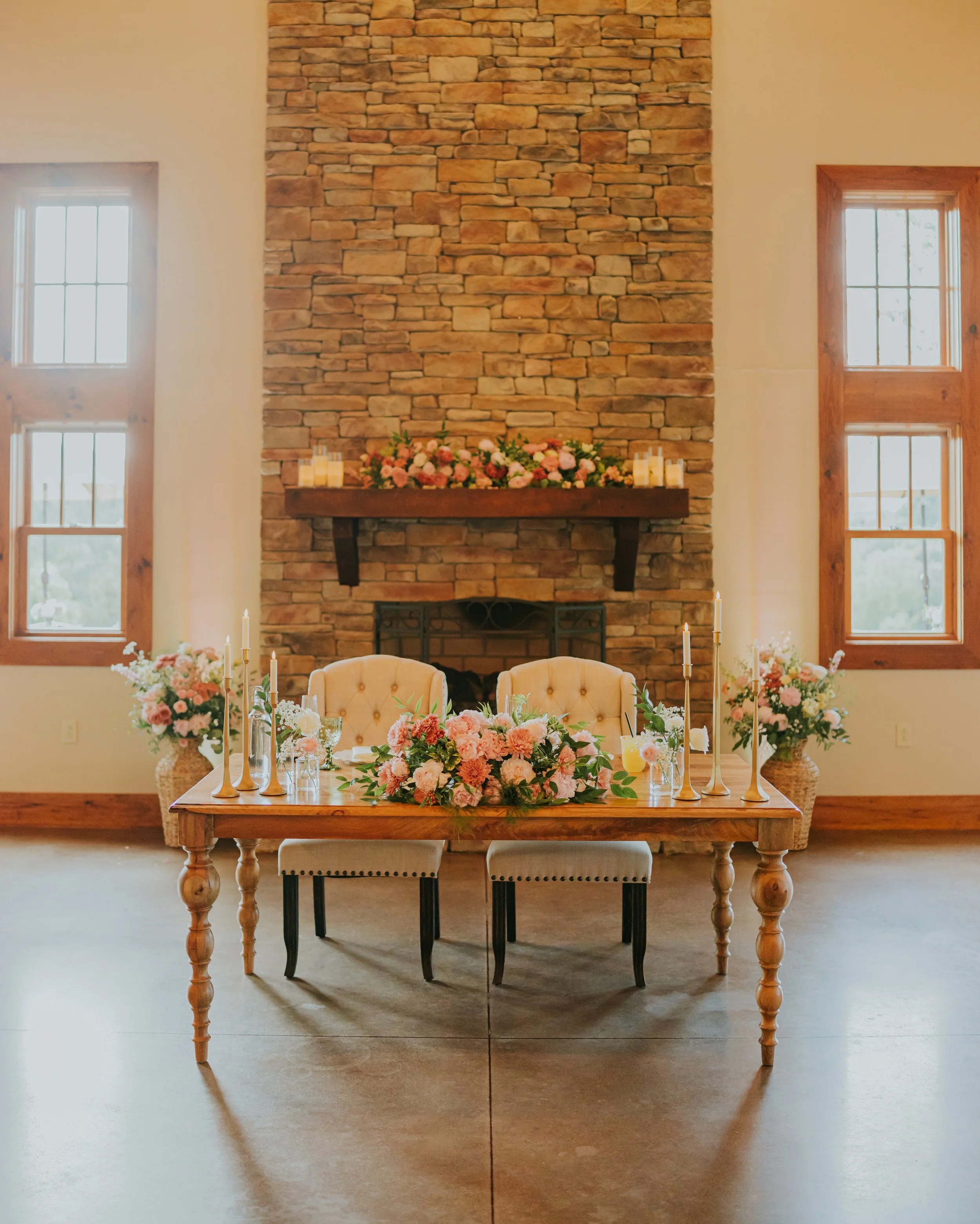 A wooden dining table decorated with pink and white flowers, surrounded by chairs, in front of a brick fireplace with floral arrangements and candles, in a room with large windows.