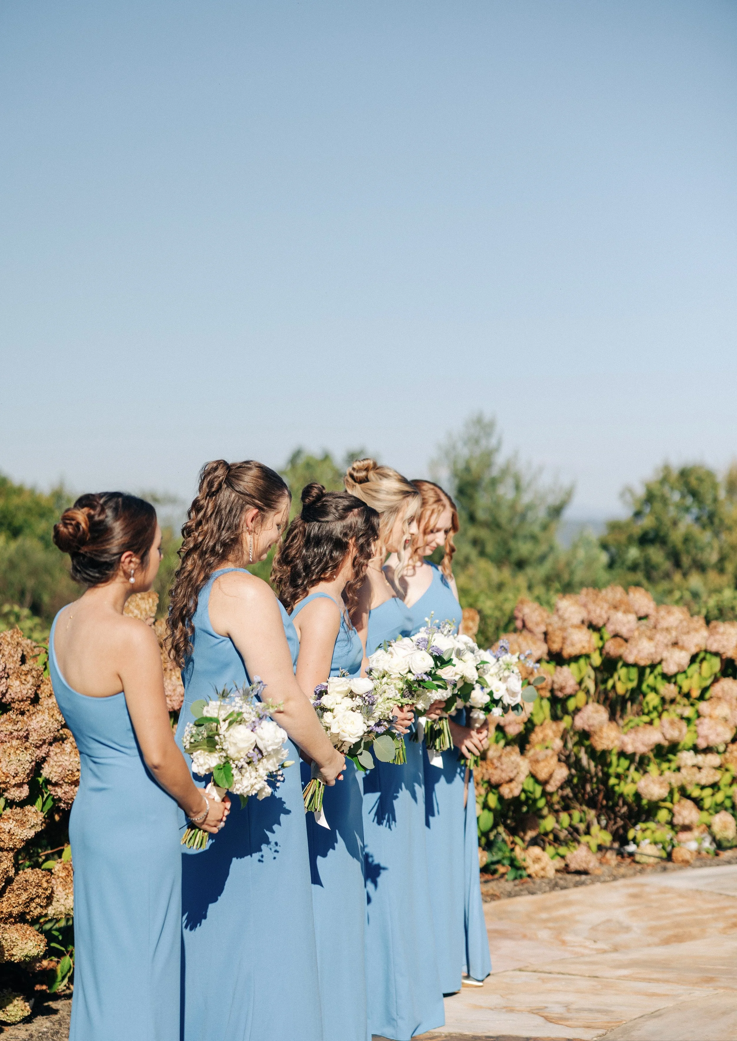 Line of women in blue dresses holding bouquets of white and purple flowers, standing outdoors with greenery and hydrangeas in the background.