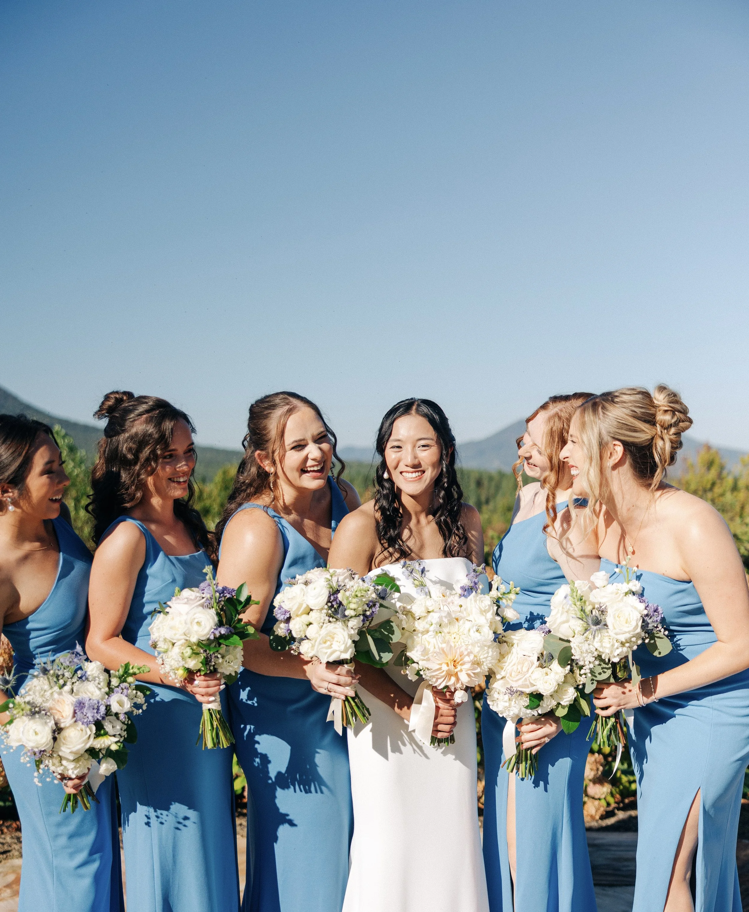 A group of women in blue dresses with a bride in white dress holding bouquets of white and purple flowers, smiling and gathered outdoors under a clear blue sky.