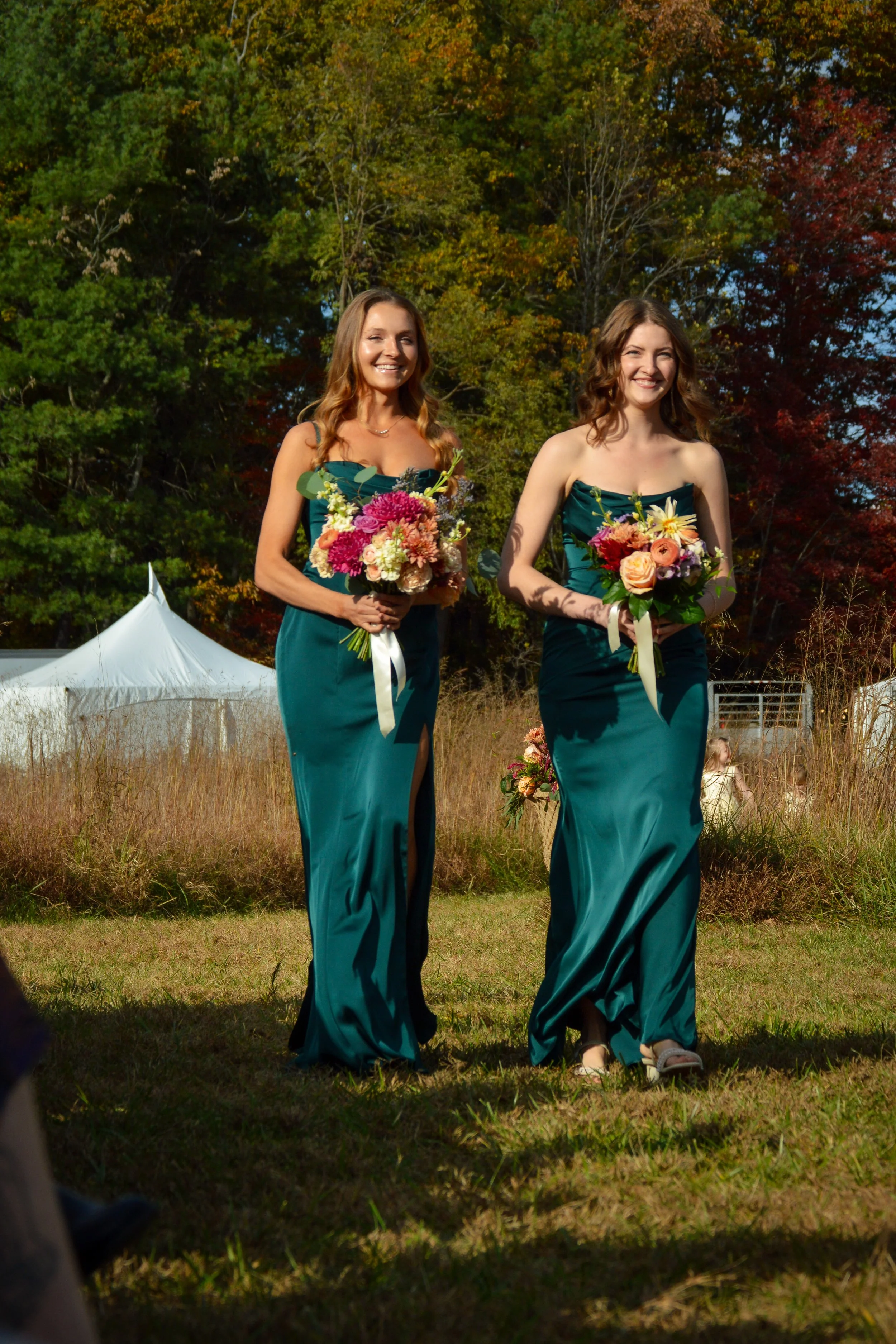 Two women in teal dresses holding bouquets of colorful flowers are walking outdoors in a grassy area with trees in autumn foliage in the background.