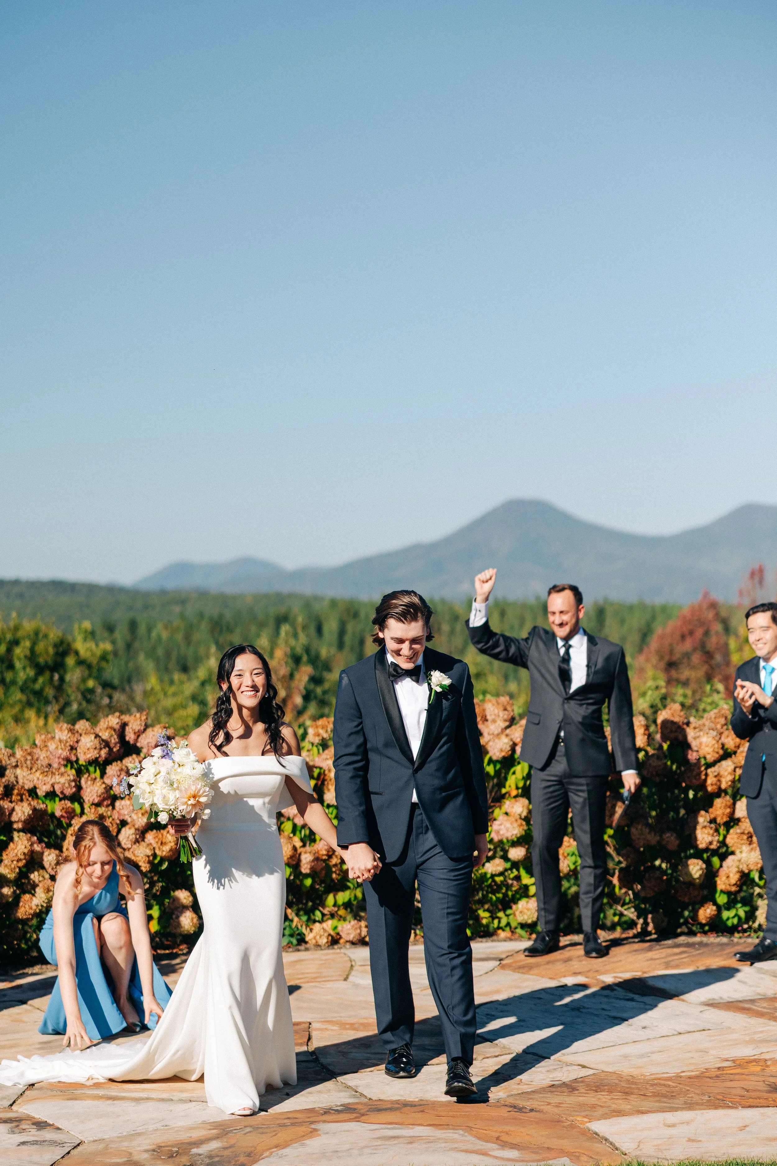 A group of wedding guests walking on a stone path outdoors on a sunny day, with mountains in the background and colorful flowers lining the path.