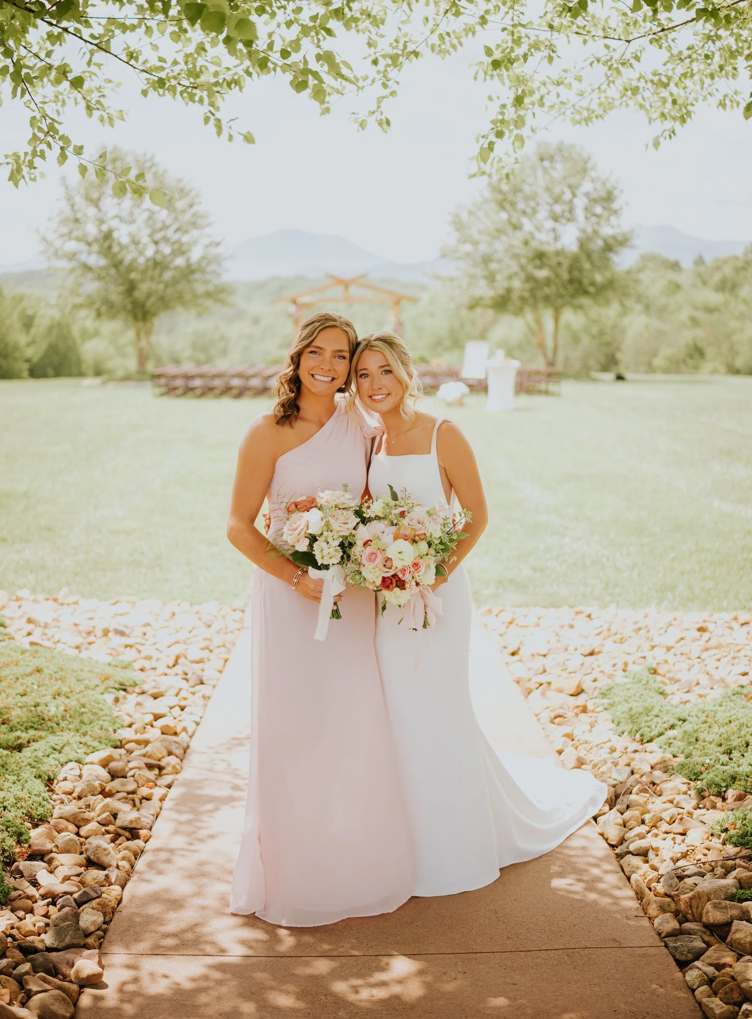 Two women on a wedding day, standing on a stone and concrete pathway outdoors, holding a bouquet of flowers, surrounded by trees and greenery, with a scenic mountain view in the background.