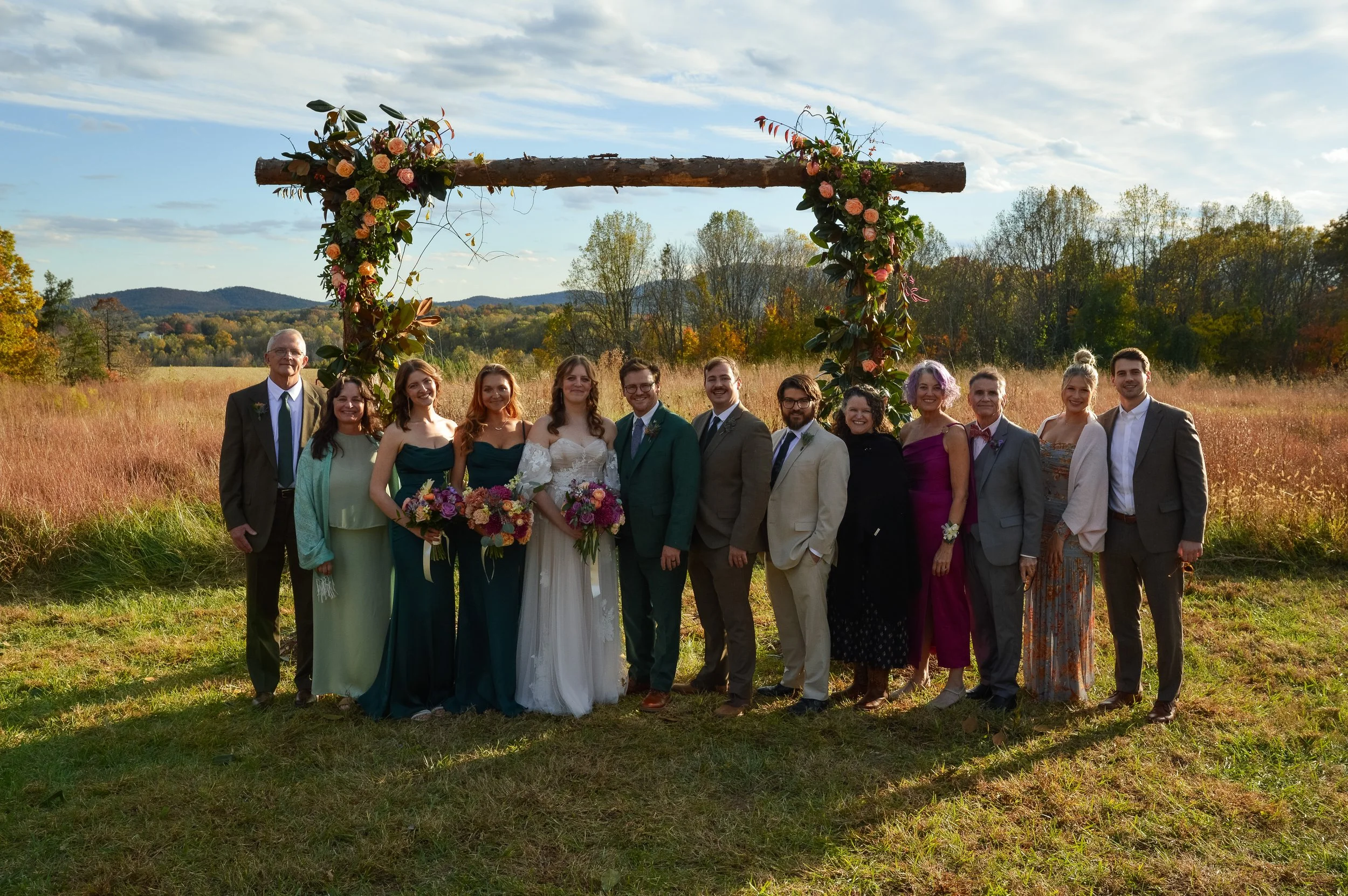 A group of people dressed in formal attire standing outdoors under a flower-decorated wooden arch on a field with trees and hills in the background.