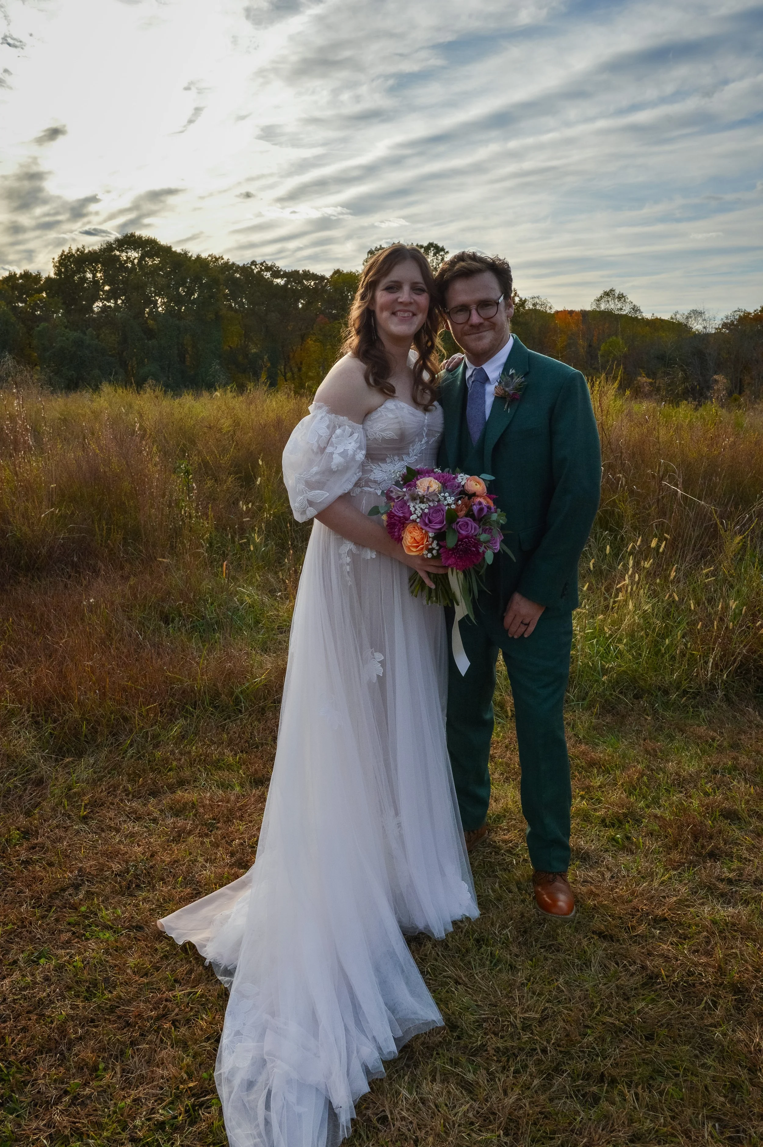 A bride and groom standing outdoors in a field at sunset, smiling and holding a colorful bouquet of flowers, with trees and a cloudy sky in the background.