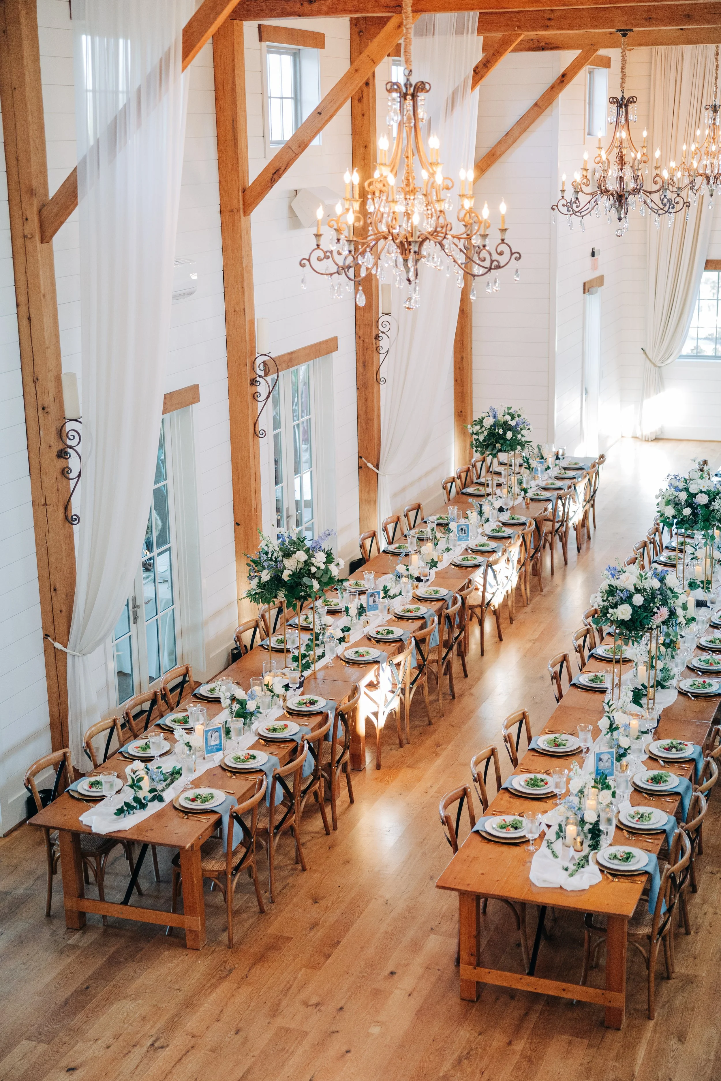 Wedding reception setup in a bright wooden hall with long tables decorated with white tablecloths, green leafy centerpieces, plates, glasses, and silverware, illuminated by large chandeliers and natural sunlight streaming through windows with white c