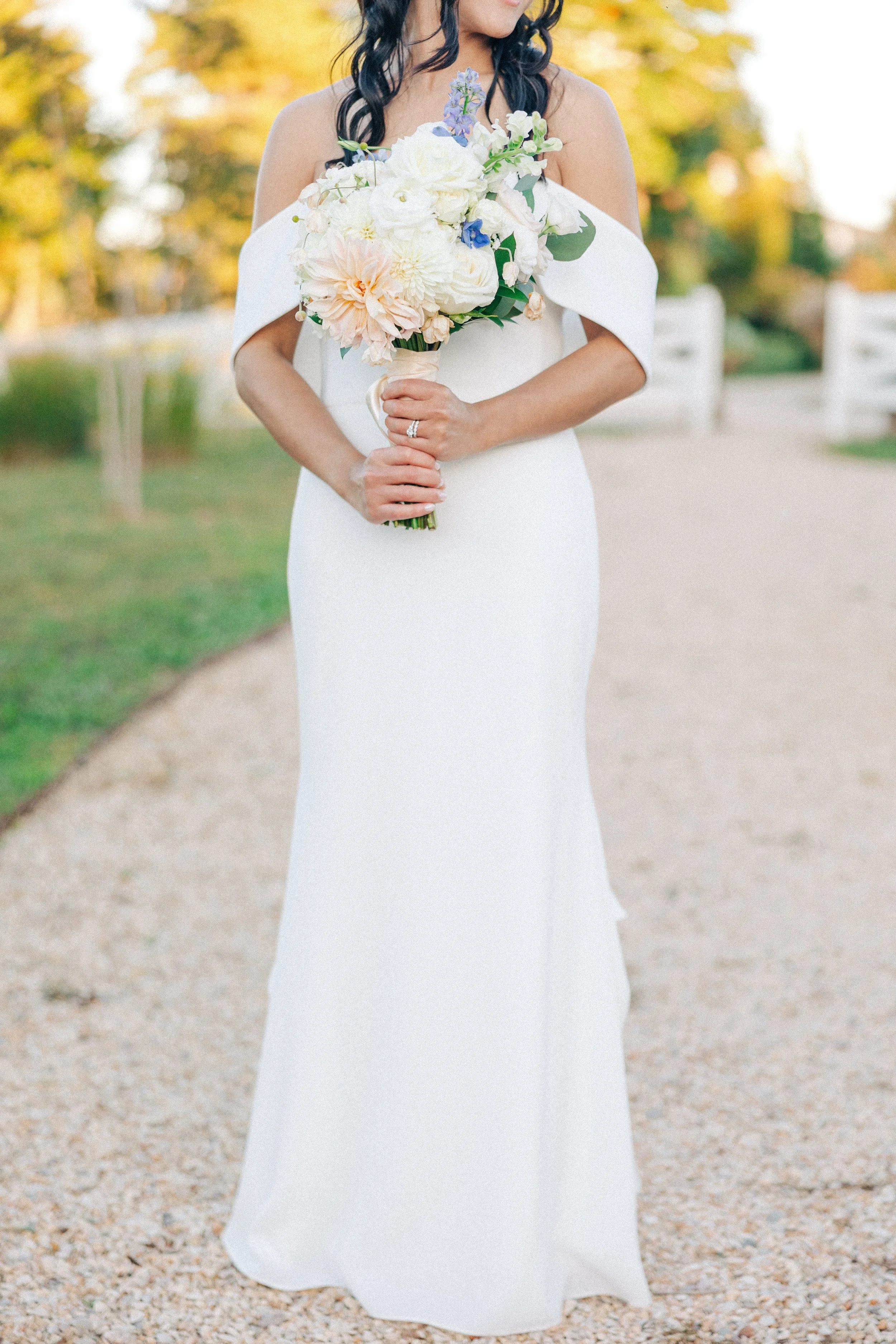 Bride in a white wedding dress holding a bouquet of white, pink, and purple flowers outdoors with autumn trees in the background.