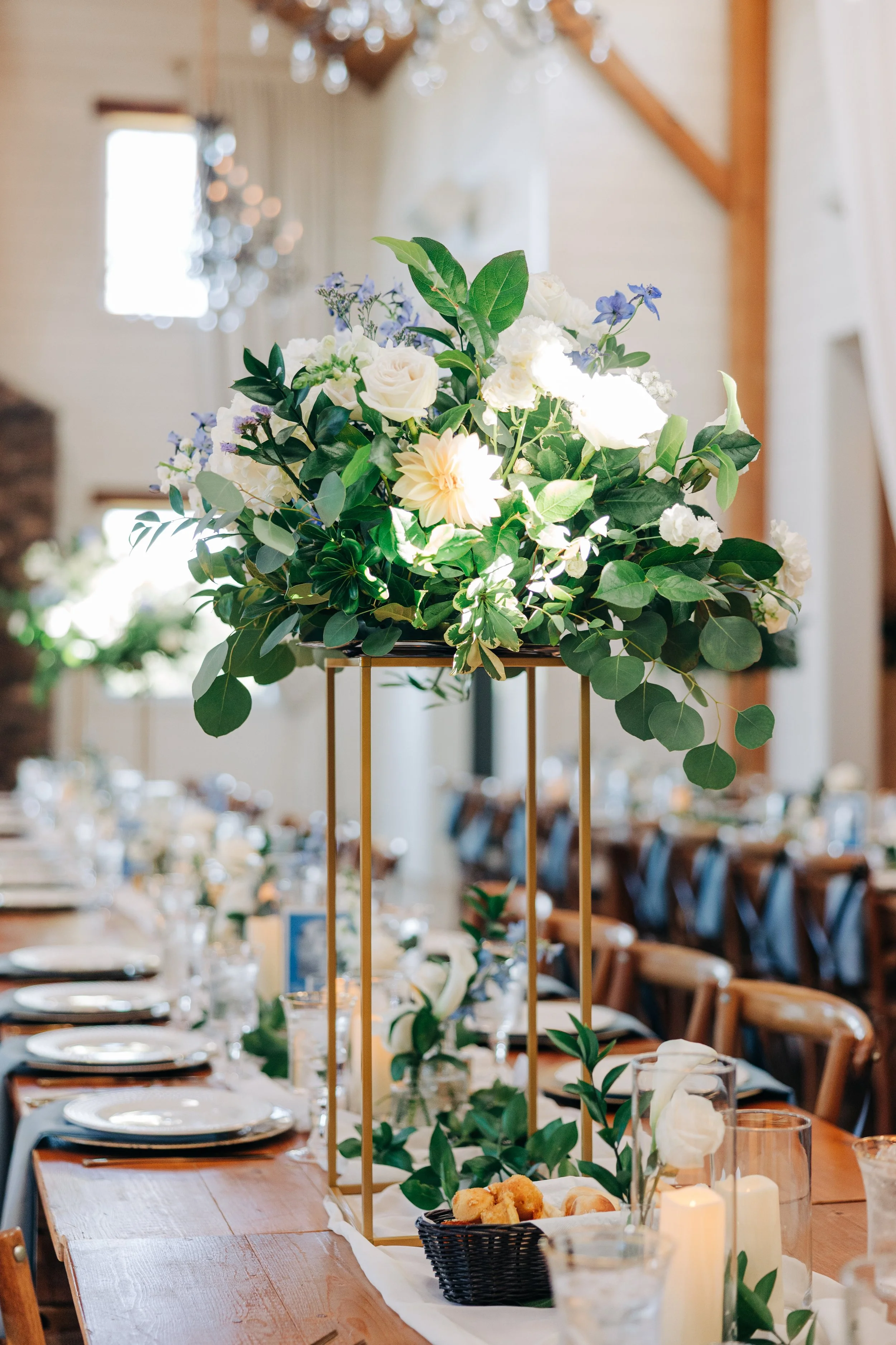 A floral centerpiece on a wedding or event table with white roses, greenery, and purple flowers, set in a tall golden frame, in a bright indoor venue with wooden beams and table settings in the background.