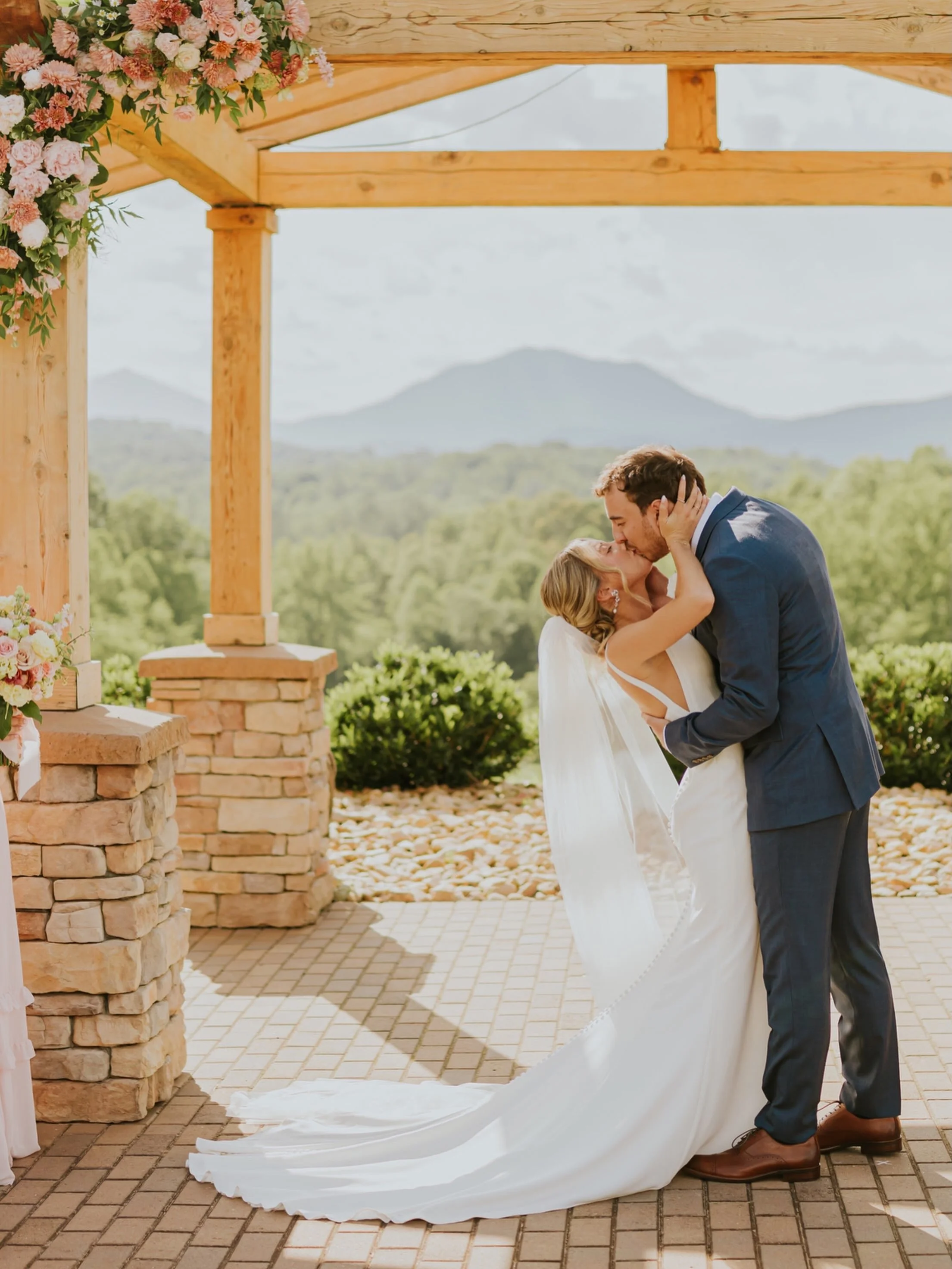 A bride and groom share a kiss under a wooden wedding arch decorated with pink and white flowers, with a scenic mountain backdrop.