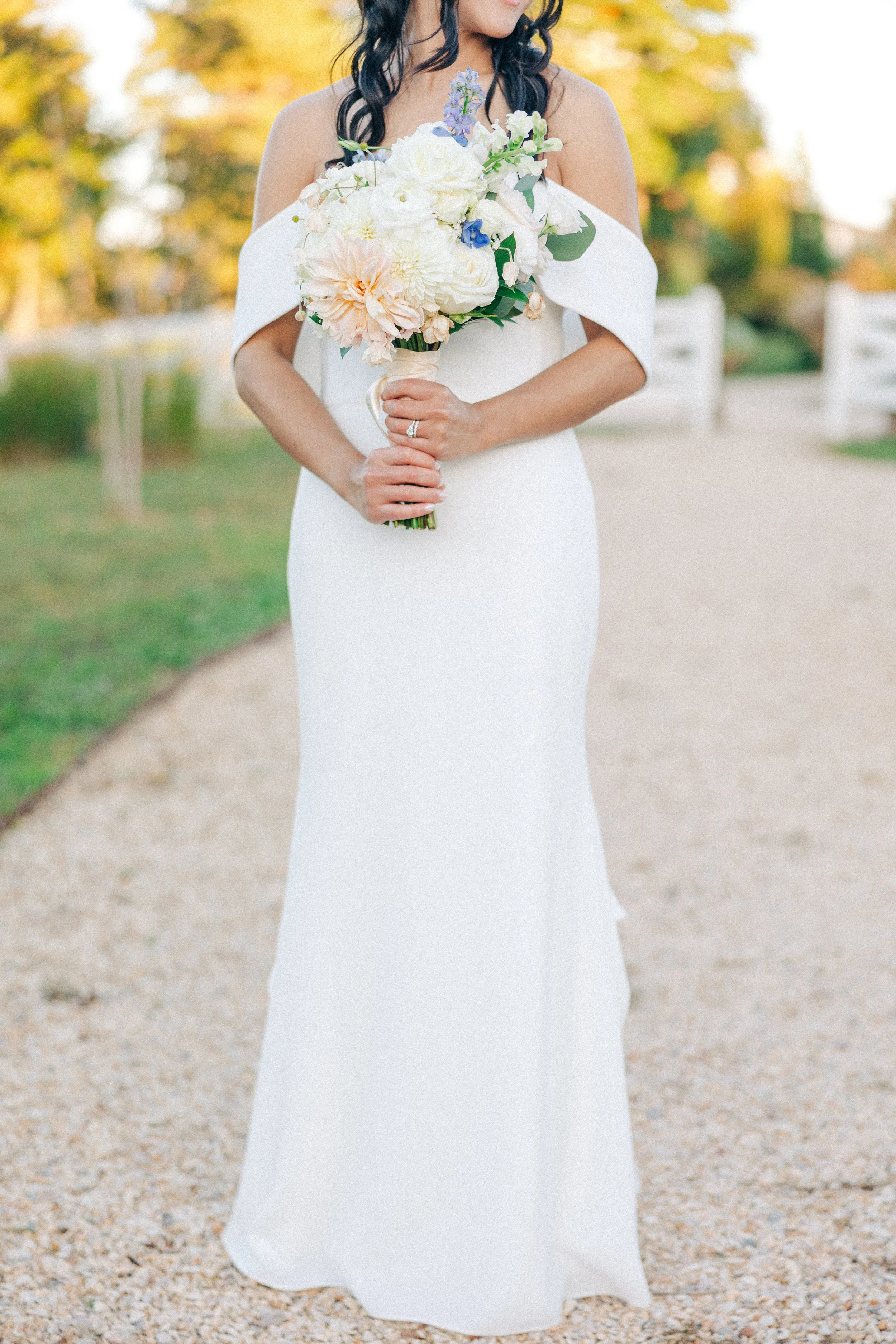 A woman in a white off-the-shoulder wedding dress holding a large bouquet of white and pastel-colored flowers, standing outdoors on a gravel path with trees in the background.