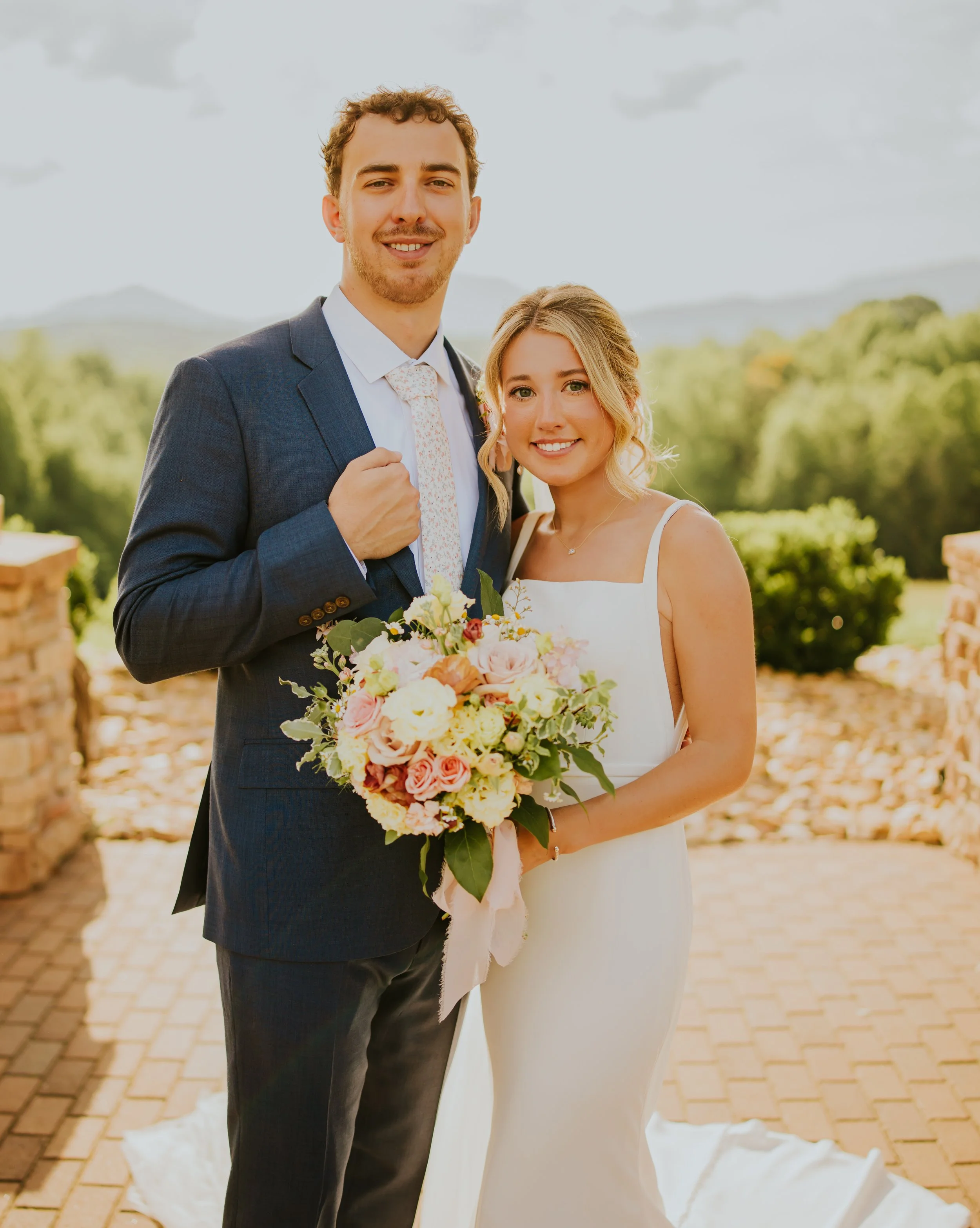 A newlywed couple outdoors at sunset, the groom in a dark suit and the bride in a white wedding dress, holding a bouquet of pink and white flowers.