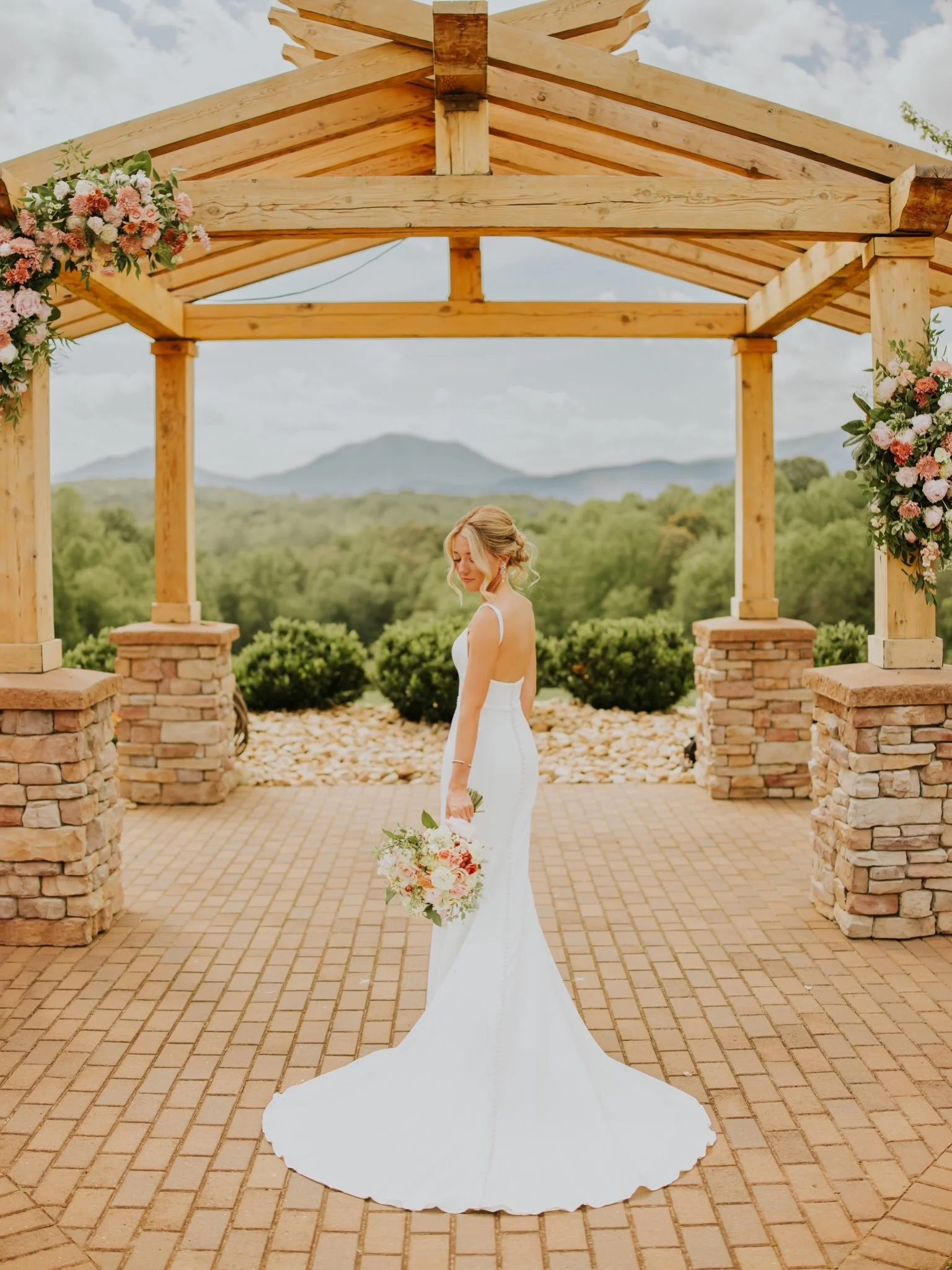 A bride in a white wedding dress standing under a wooden arch decorated with pink and white flowers against a scenic outdoor landscape with mountains in the background.