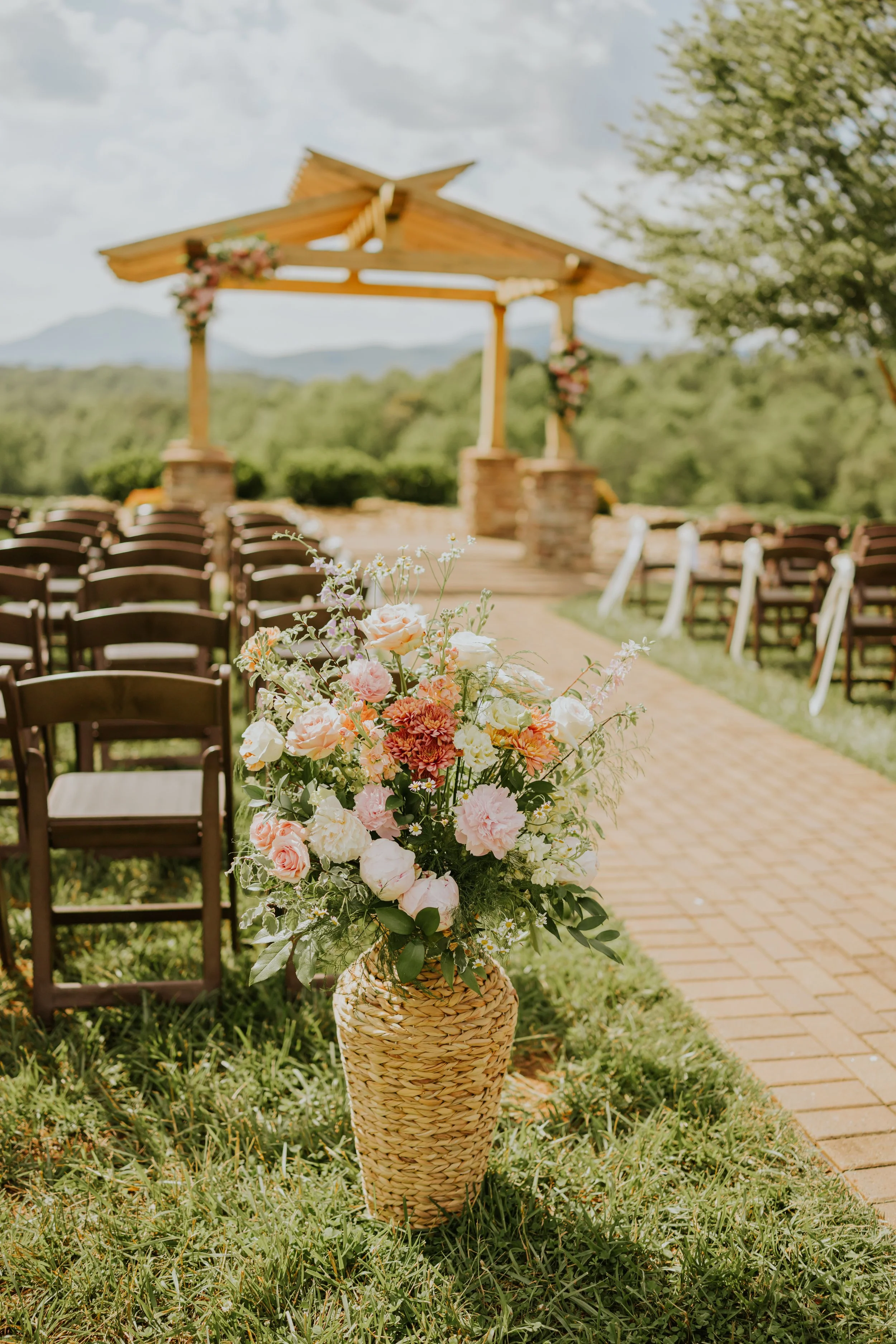 A flower arrangement in a woven vase at an outdoor wedding ceremony with wooden chairs, an arch decorated with flowers, and scenic mountains in the background.