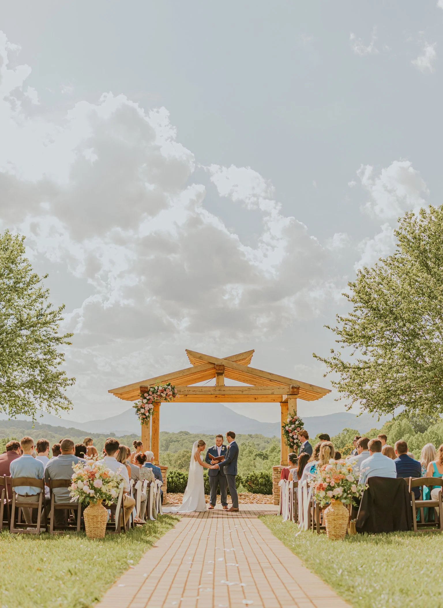 An outdoor wedding ceremony with a wooden arch decorated with flowers, set in a scenic area with mountains in the background. The bride and groom are standing under the arch, facing each other, with guests seated on either side.