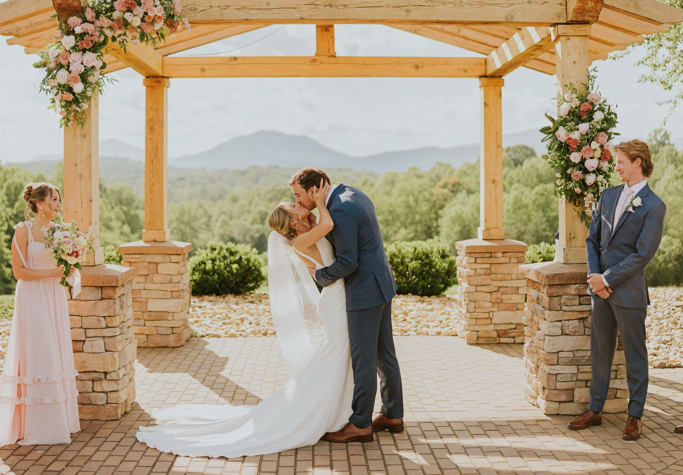 A wedding ceremony taking place outdoors under a wooden pergola decorated with pink and white flowers, with the bride and groom kissing in the center. The bride is wearing a white wedding dress and the groom is in a dark suit. A bridesmaid holding a 