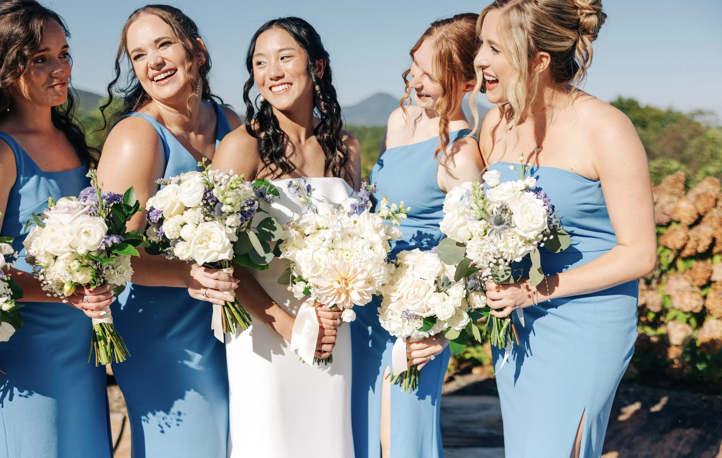 A group of women dressed in blue bridesmaid dresses holding white and purple flower bouquets, standing outdoors at a wedding.