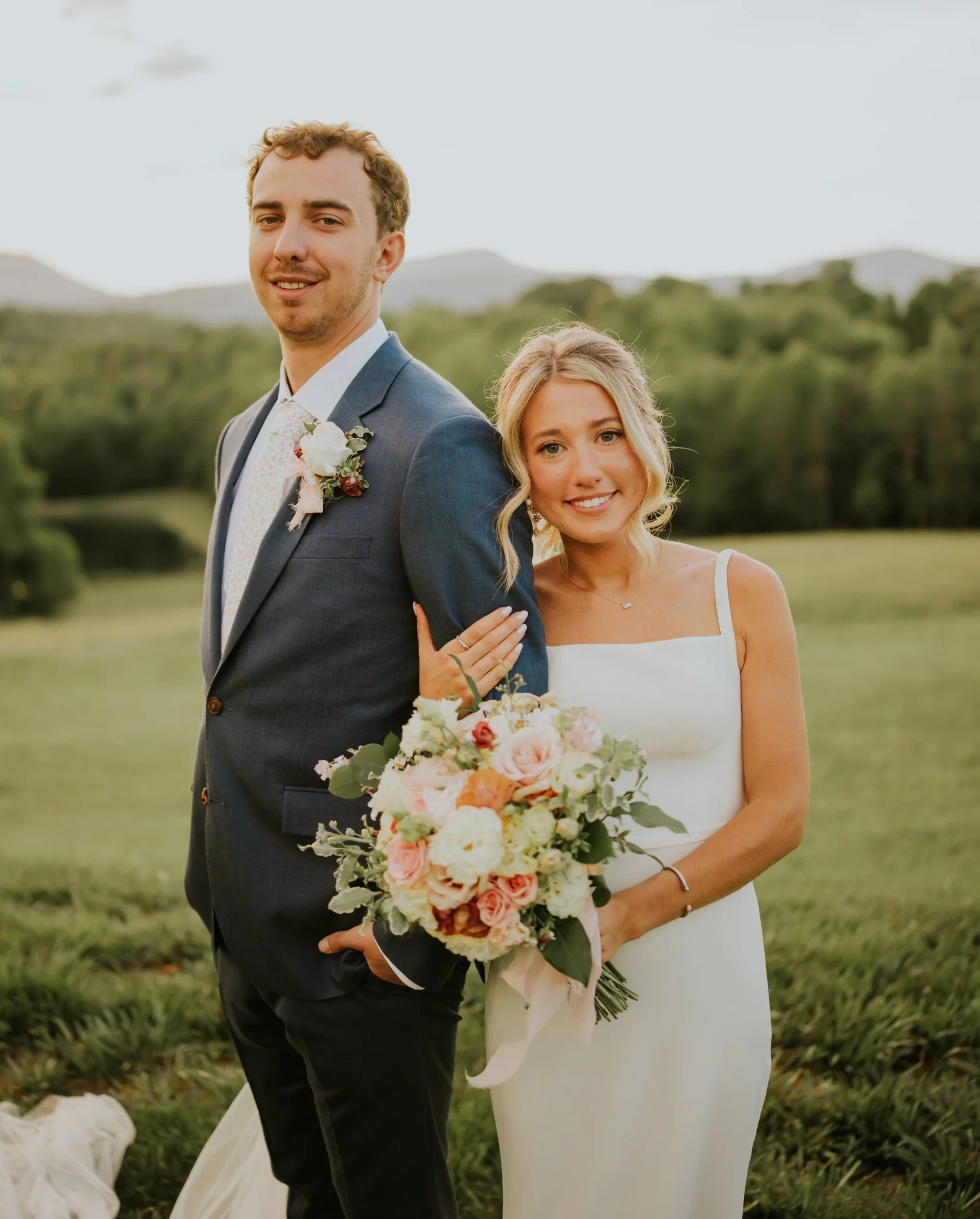 A smiling man in a suit and woman in a white dress standing outdoors in a field with trees and mountains in the background, holding a bouquet of flowers, possibly during a wedding or engagement photo.