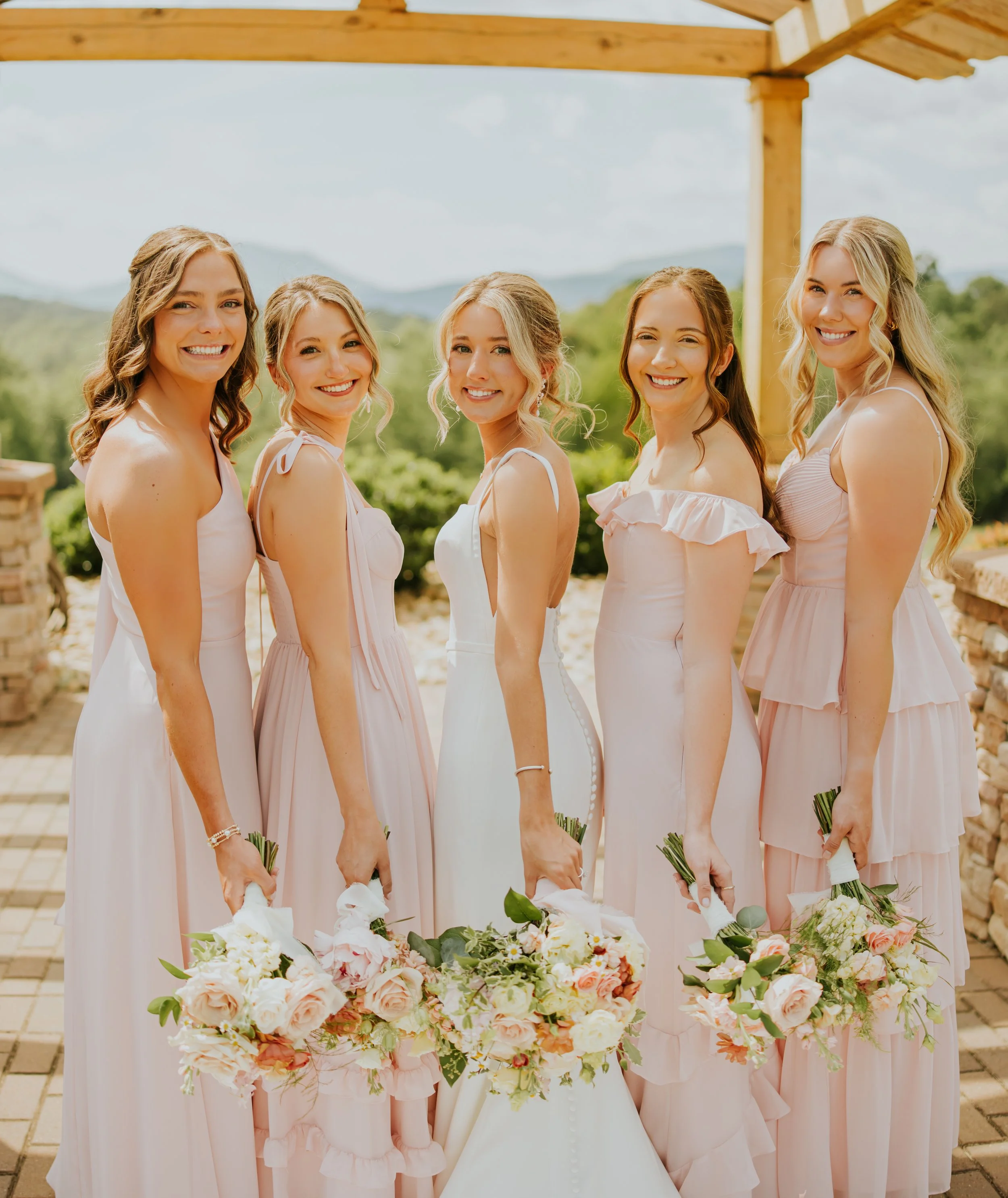 Group of six women in pastel dresses holding bouquets of flowers, standing outdoors under a wooden pergola with a scenic mountain backdrop.