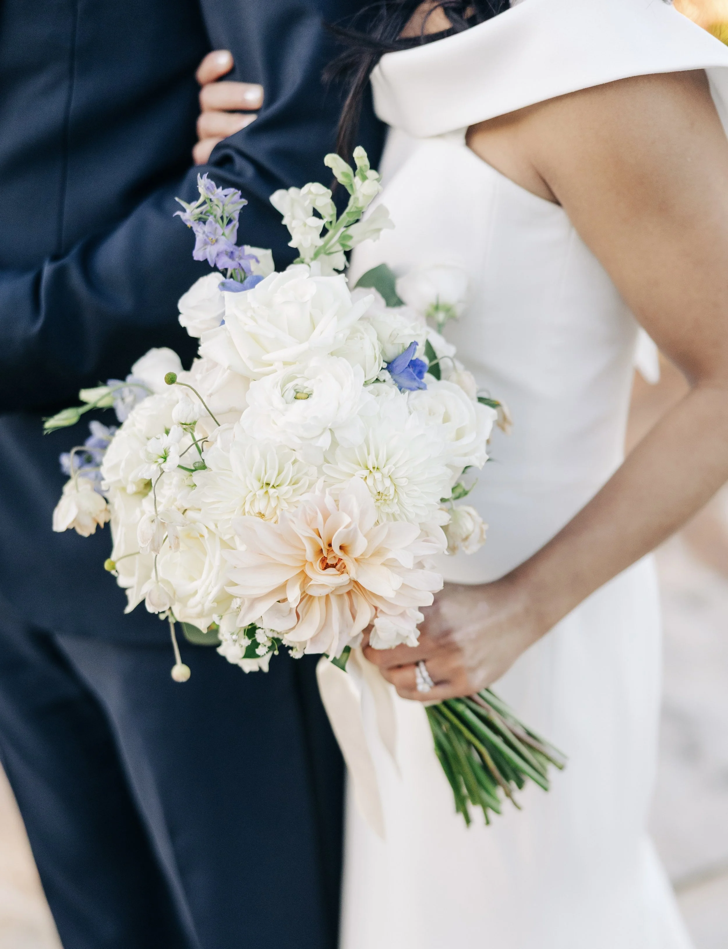 Close-up of a bride holding a bouquet of white, purple, and peach flowers, with a groom in a dark suit beside her, during a wedding ceremony.