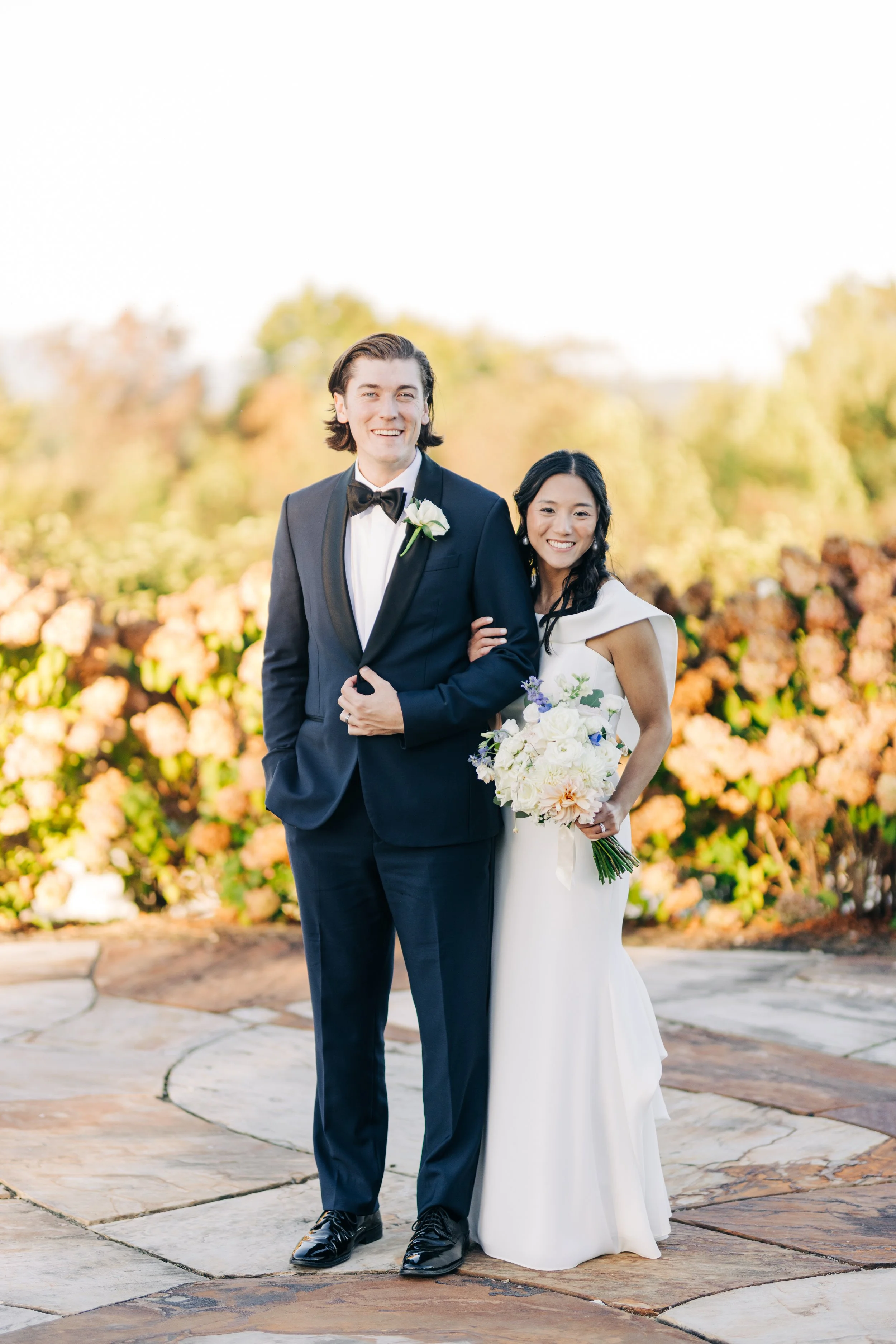 A smiling couple dressed in wedding attire standing outdoors with trees and flowers in the background during daytime. The groom wears a black tuxedo with a bow tie and a white boutonniere, while the bride wears a white wedding gown and holds a bouque