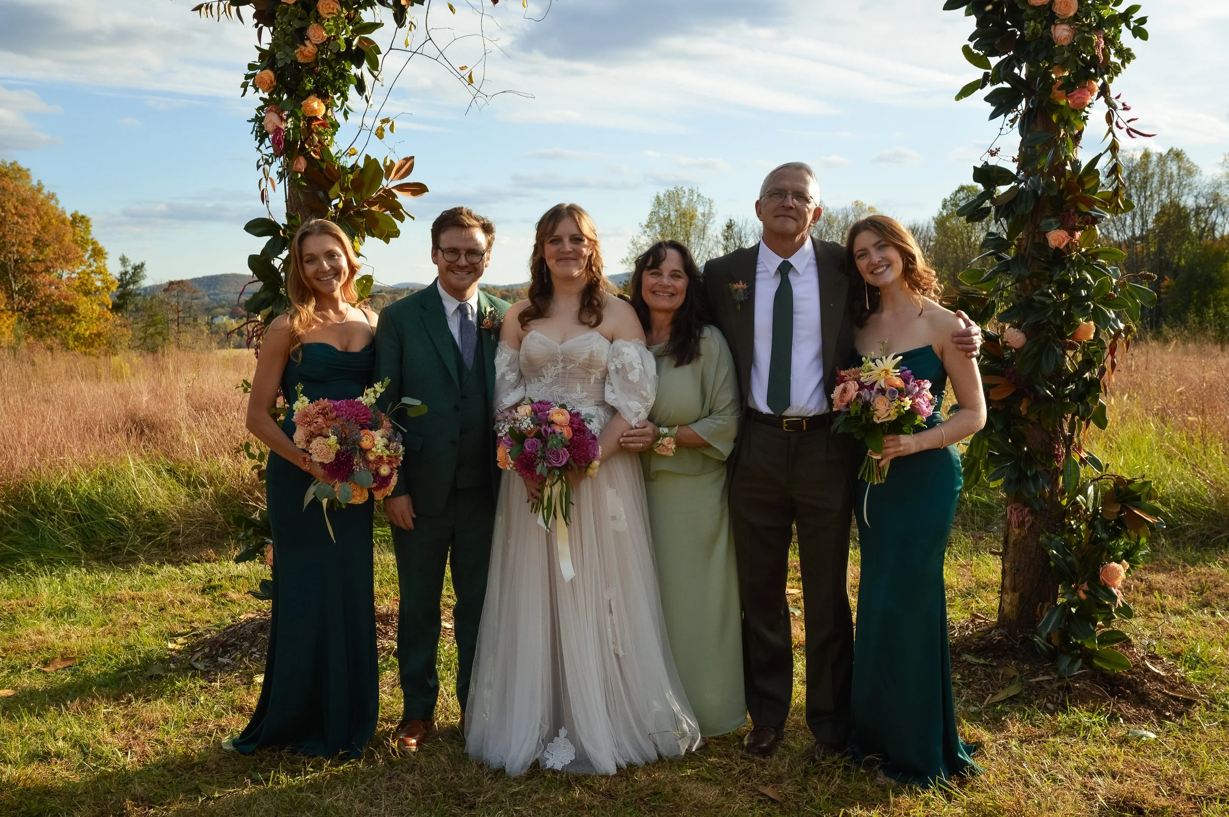 A group of seven people standing outdoors under a floral arch, smiling at the camera, during a wedding celebration. The bride is dressed in a white wedding gown holding a bouquet, with two bridesmaids in matching teal dresses on either side. Two men 