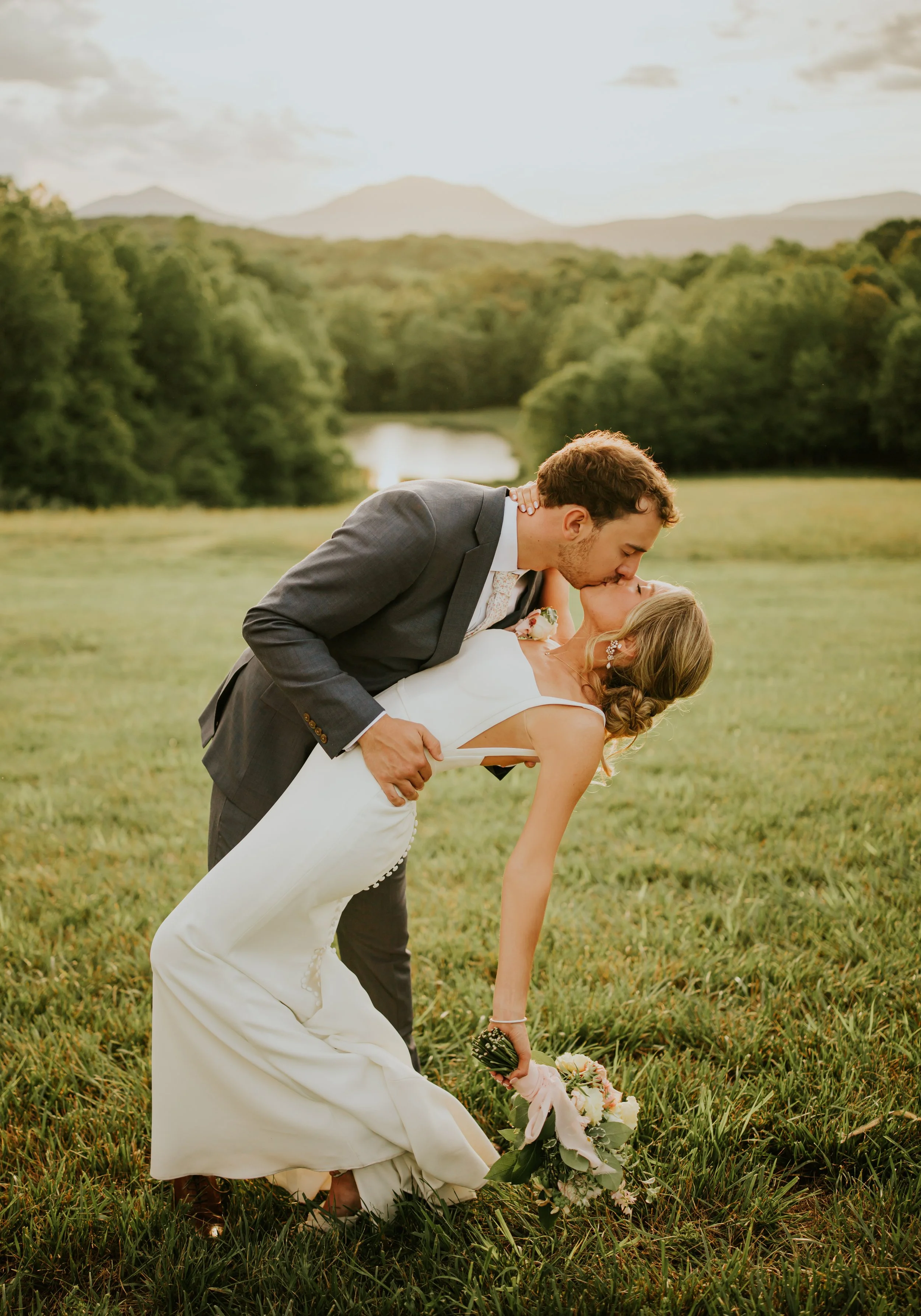 A newlywed couple sharing a kiss outdoors in a field with mountains and a lake in the background during sunset.