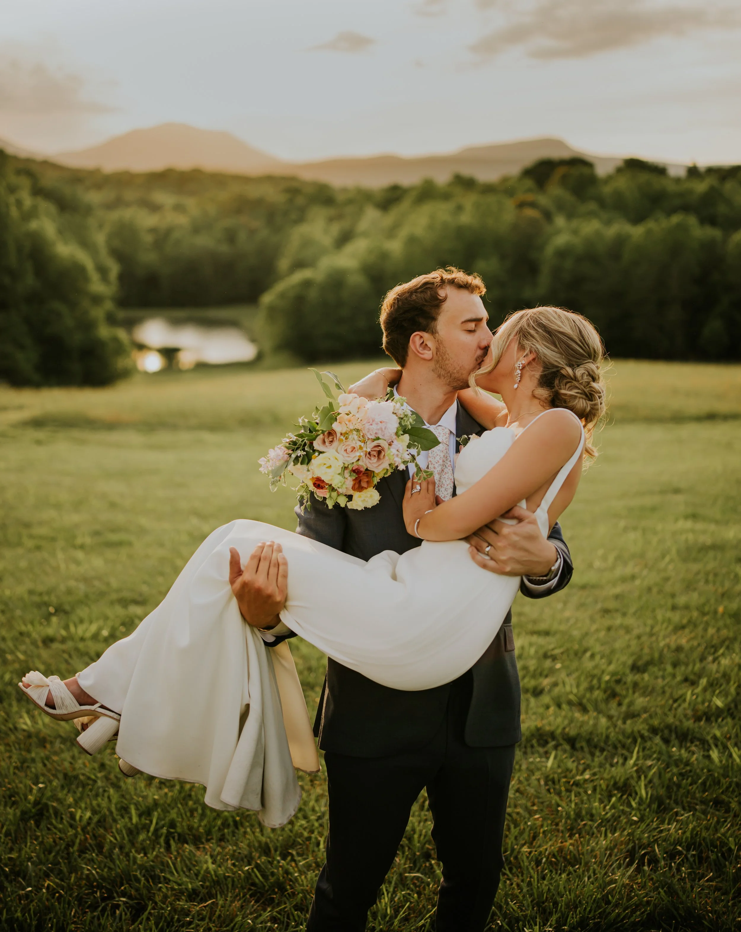 A groom in a dark suit holding and kissing a bride in a white wedding dress outdoors in a green field, with a mountain range and cloudy sky in the background, during sunset.