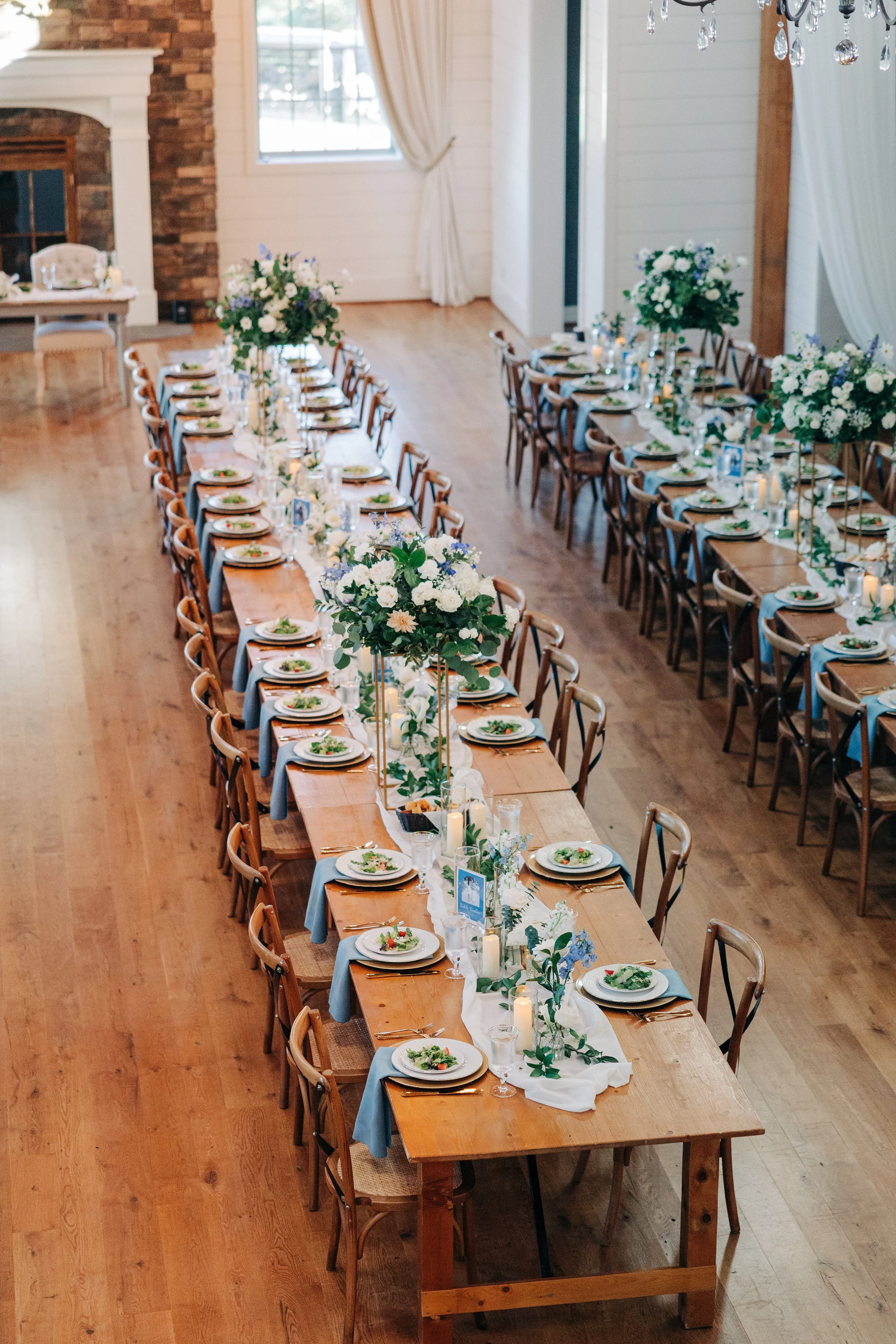 Wedding reception table set with plates, glasses, floral centerpieces, and candles in a rustic venue with wooden floors and white walls.