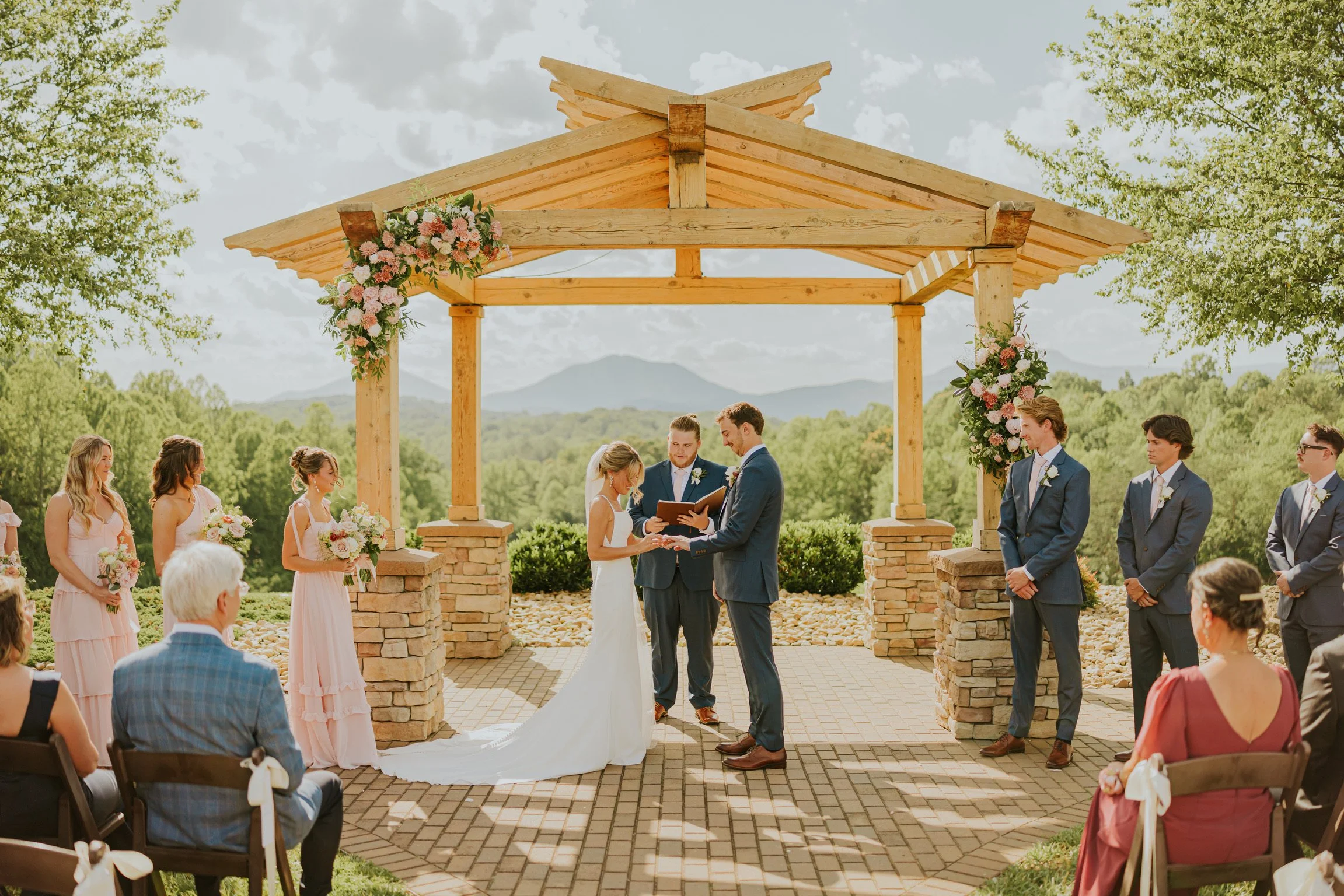 Outdoor wedding ceremony with a wooden arch decorated with pink and white flowers, couple exchanging vows, and guests seated on either side, set in a scenic area with green trees and mountains in the background.