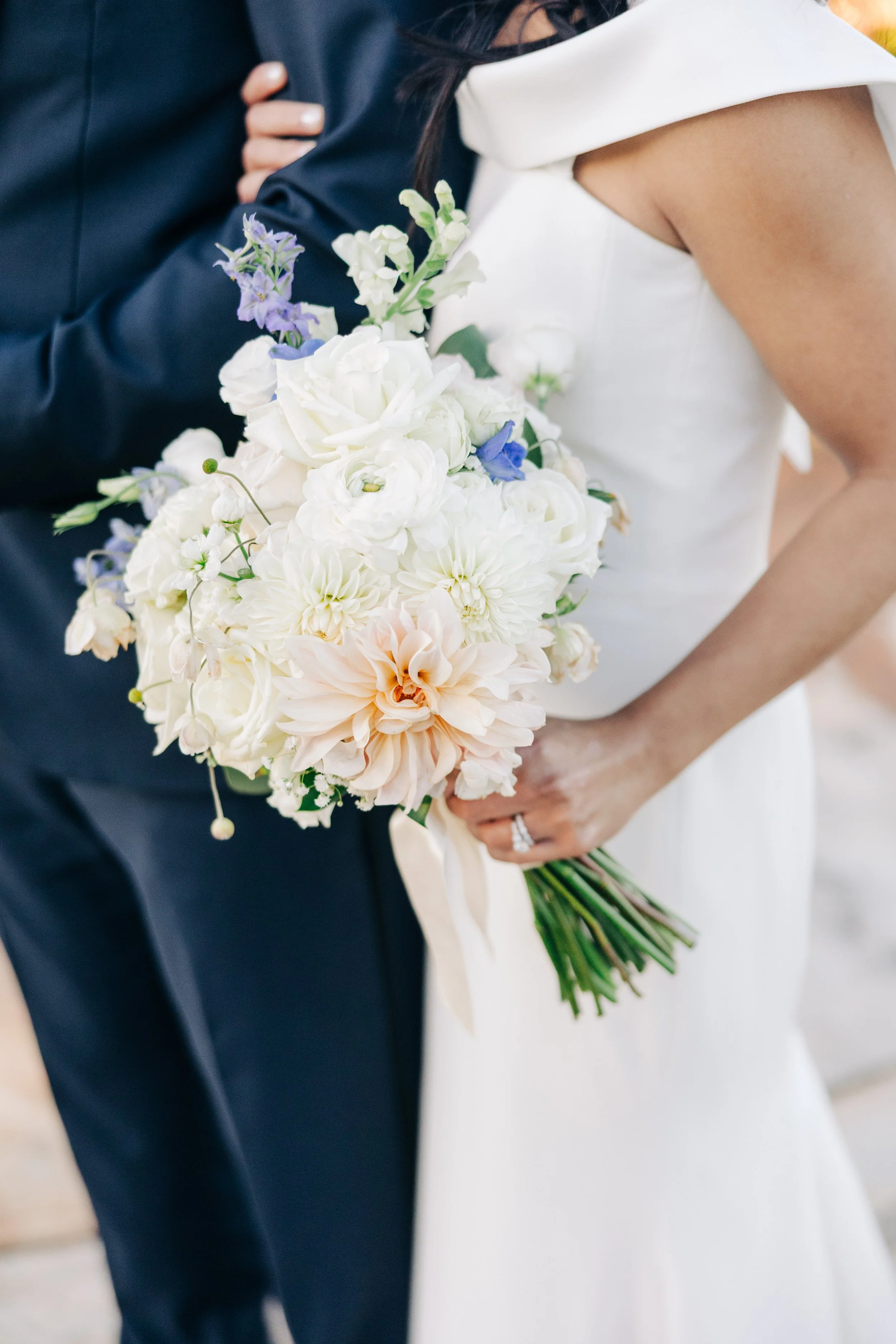 A bride in a white dress holding a bouquet of white and light pink flowers with some purple accents, standing next to a groom in a dark suit. The bride's hand, with an engagement ring and wedding band, is visible.