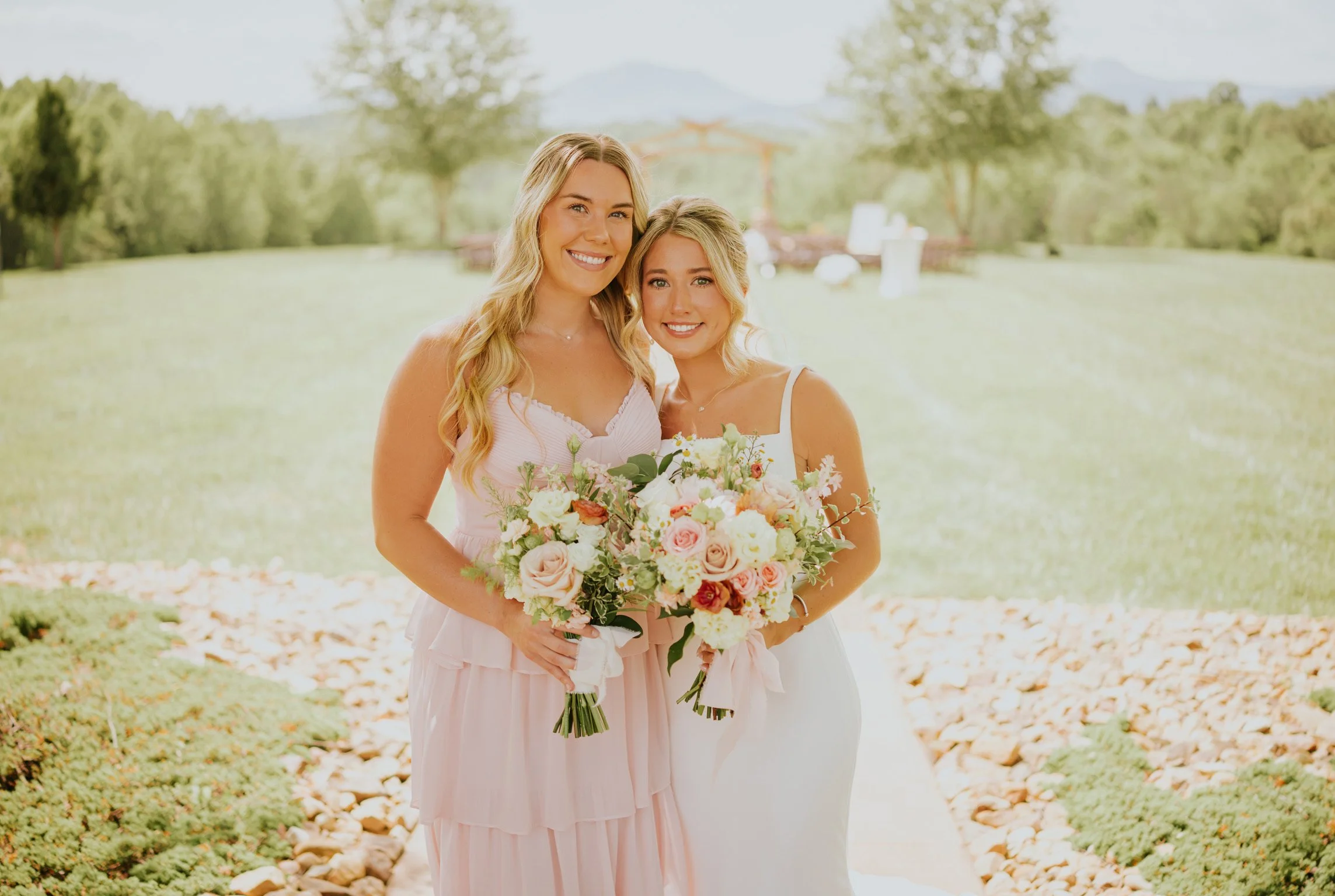 Two women, one in a pink dress and the other in a white dress, standing outdoors on a sunny day, holding a large bouquet of pink and white flowers, smiling.