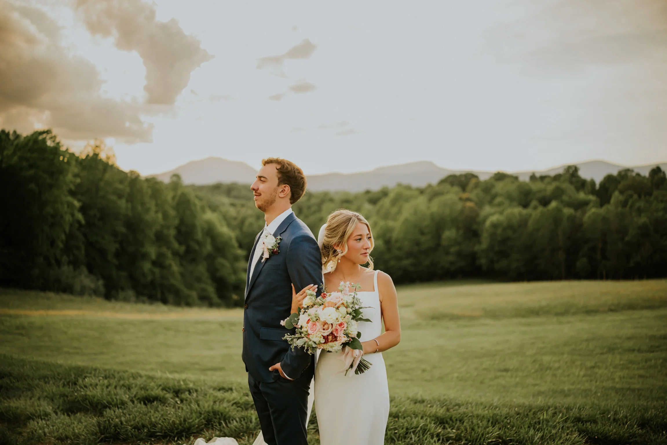 A bride and groom standing outdoors on a grassy field with trees and mountains in the background, during sunset. The groom is dressed in a blue suit with a flower boutonniere, and the bride is in a white wedding dress holding a bouquet of flowers. Th