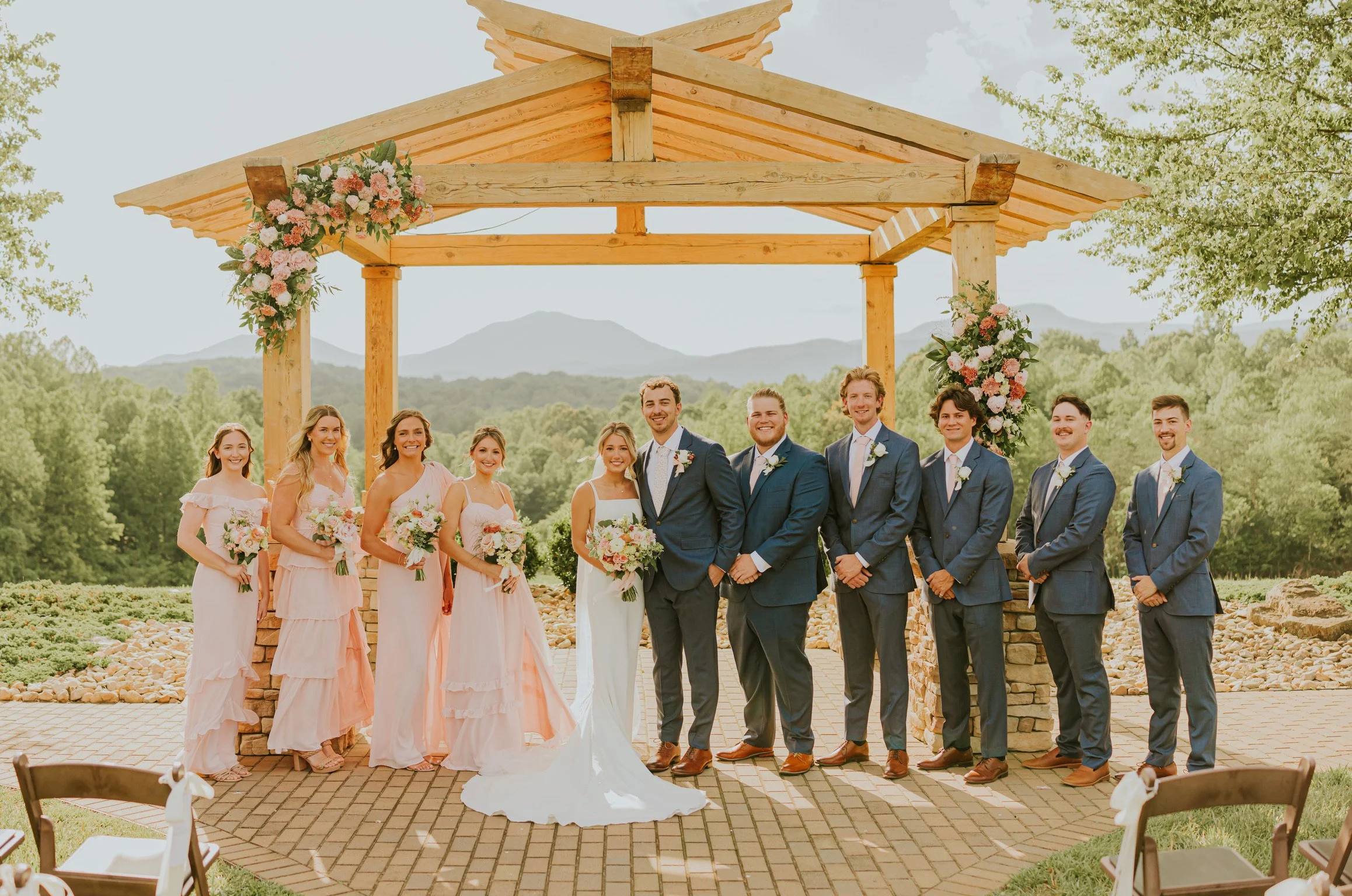 Group of wedding celebration with bride and groom in center, bridesmaids in pink dresses, groomsmen in blue suits, standing under a wooden wedding arch decorated with pink and white flowers, outdoors in a scenic mountainous area.