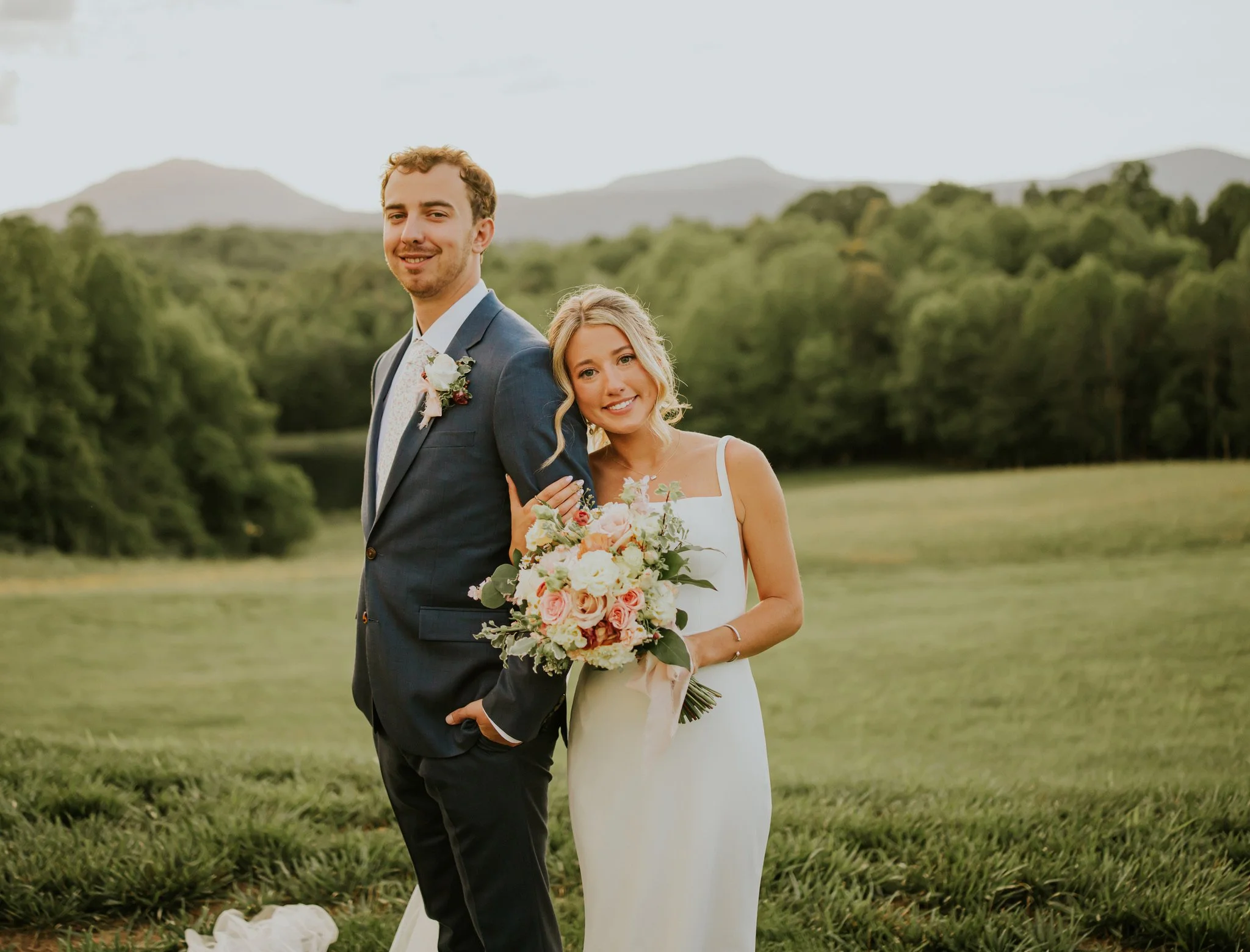 A bride and groom standing together outdoors during a wedding ceremony, with trees and mountains in the background. The groom is wearing a dark suit with a white shirt and a boutonniere, and the bride is wearing a white dress, holding a large bouquet