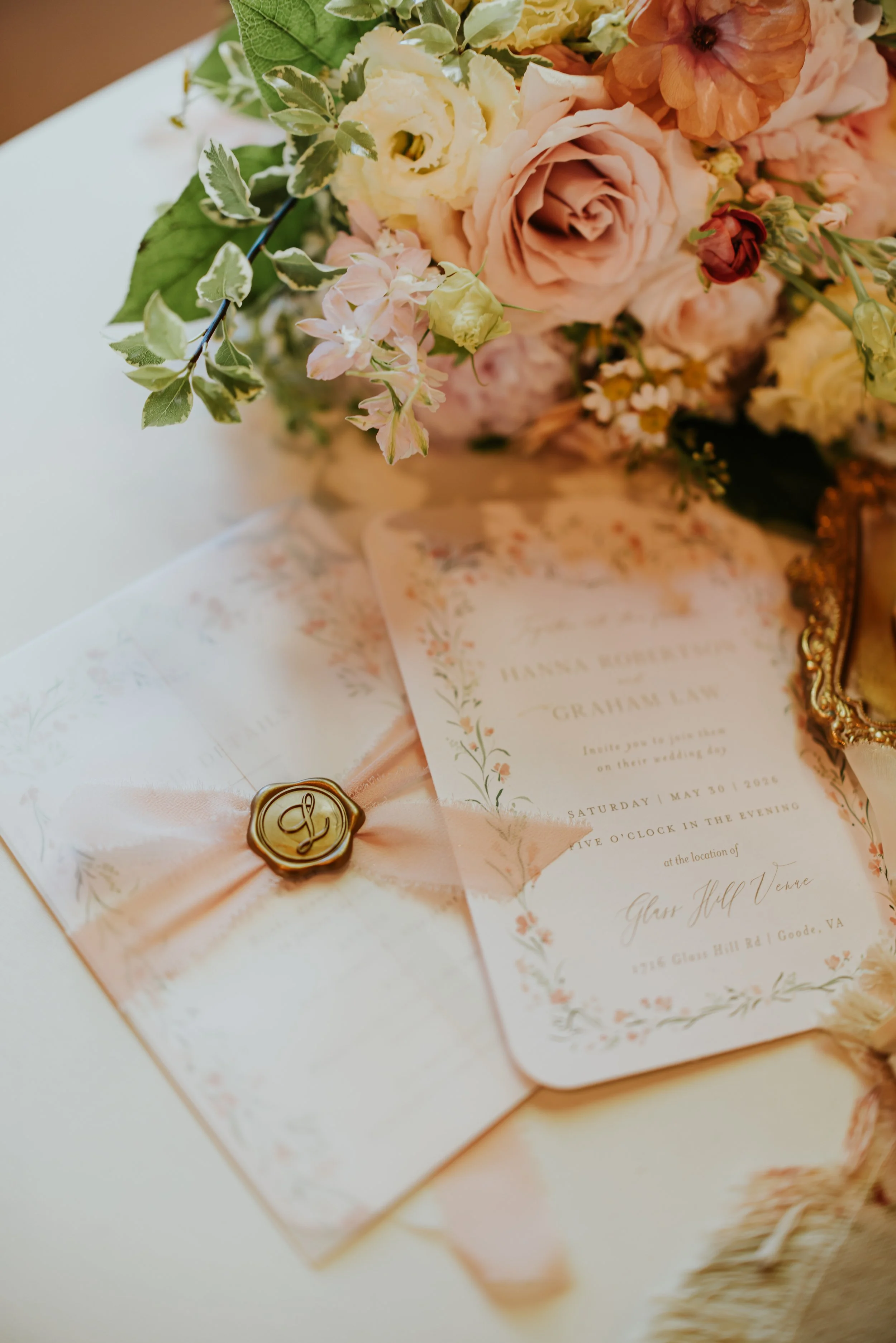 A floral arrangement with peach, cream, and pink flowers and green foliage, a wedding invitation, and a pink ribbon with a gold wax seal resting on a soft fabric surface.