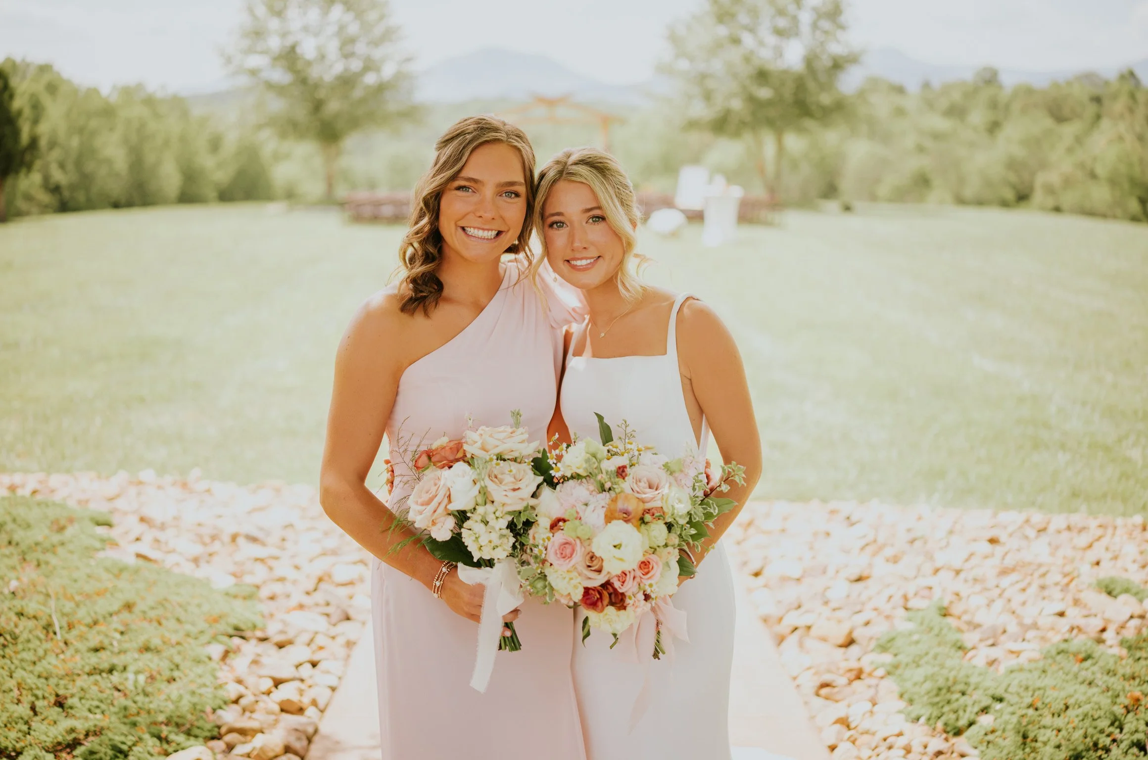 Two women in wedding dresses holding a bouquet of pink and white flowers outdoors, with trees and a lake in the background.