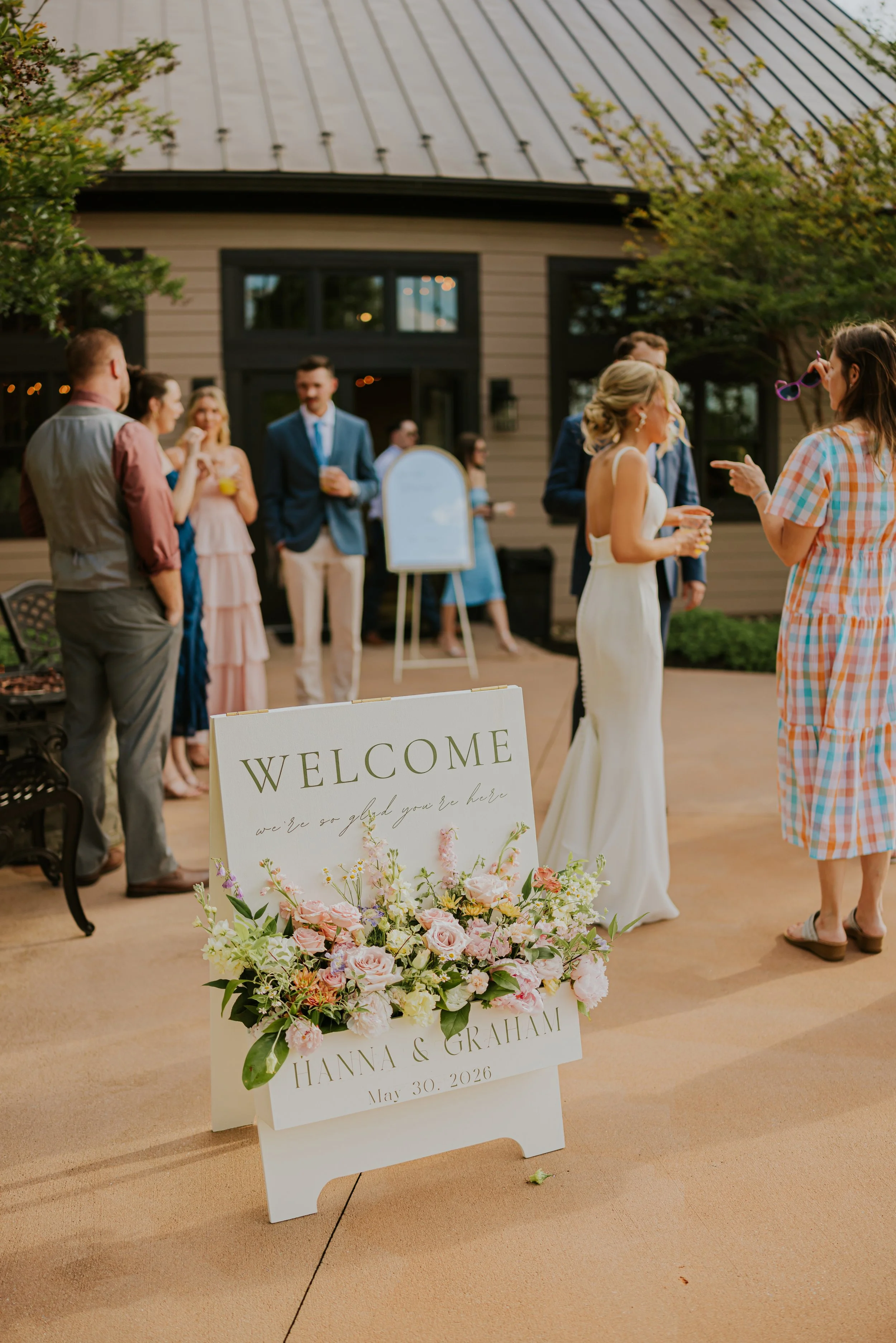 Wedding reception with guests in formal attire, floral arrangements, and welcoming sign for Hannah and Graham's wedding on May 30, 2026, outside a building with trees.