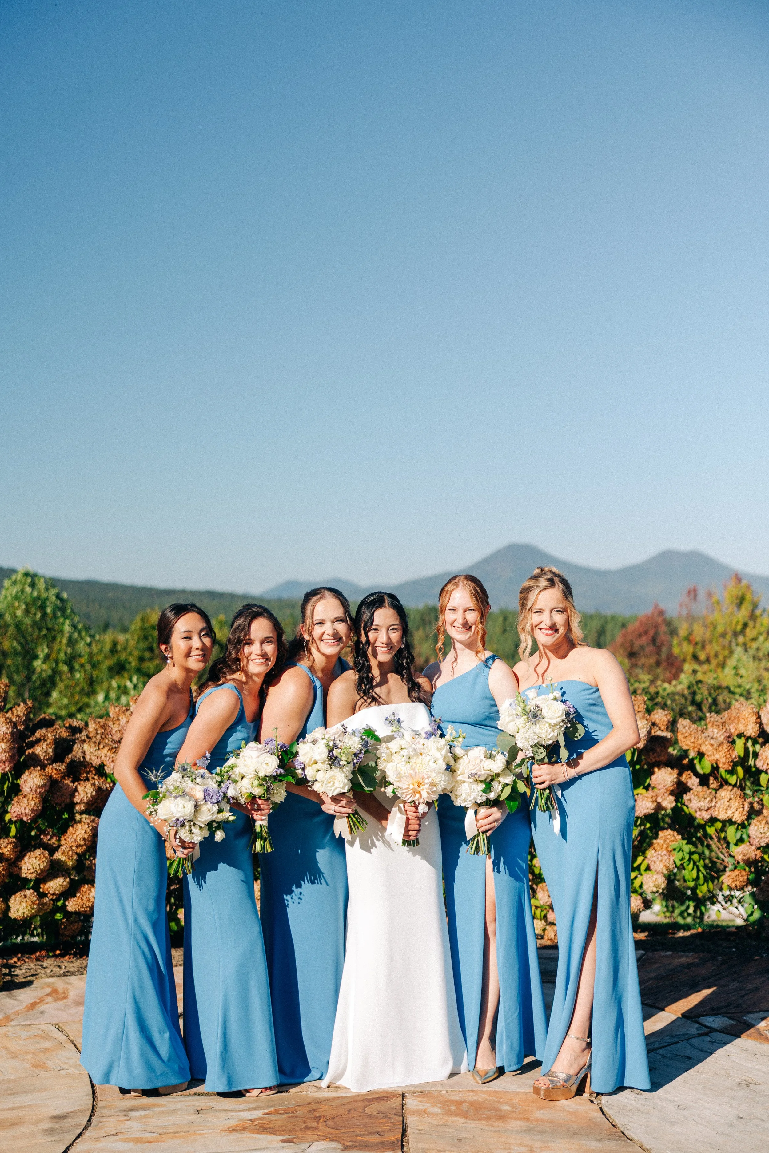 A group of women in blue dresses, holding bouquets, posed outdoors on a sunny day with a mountain in the background.