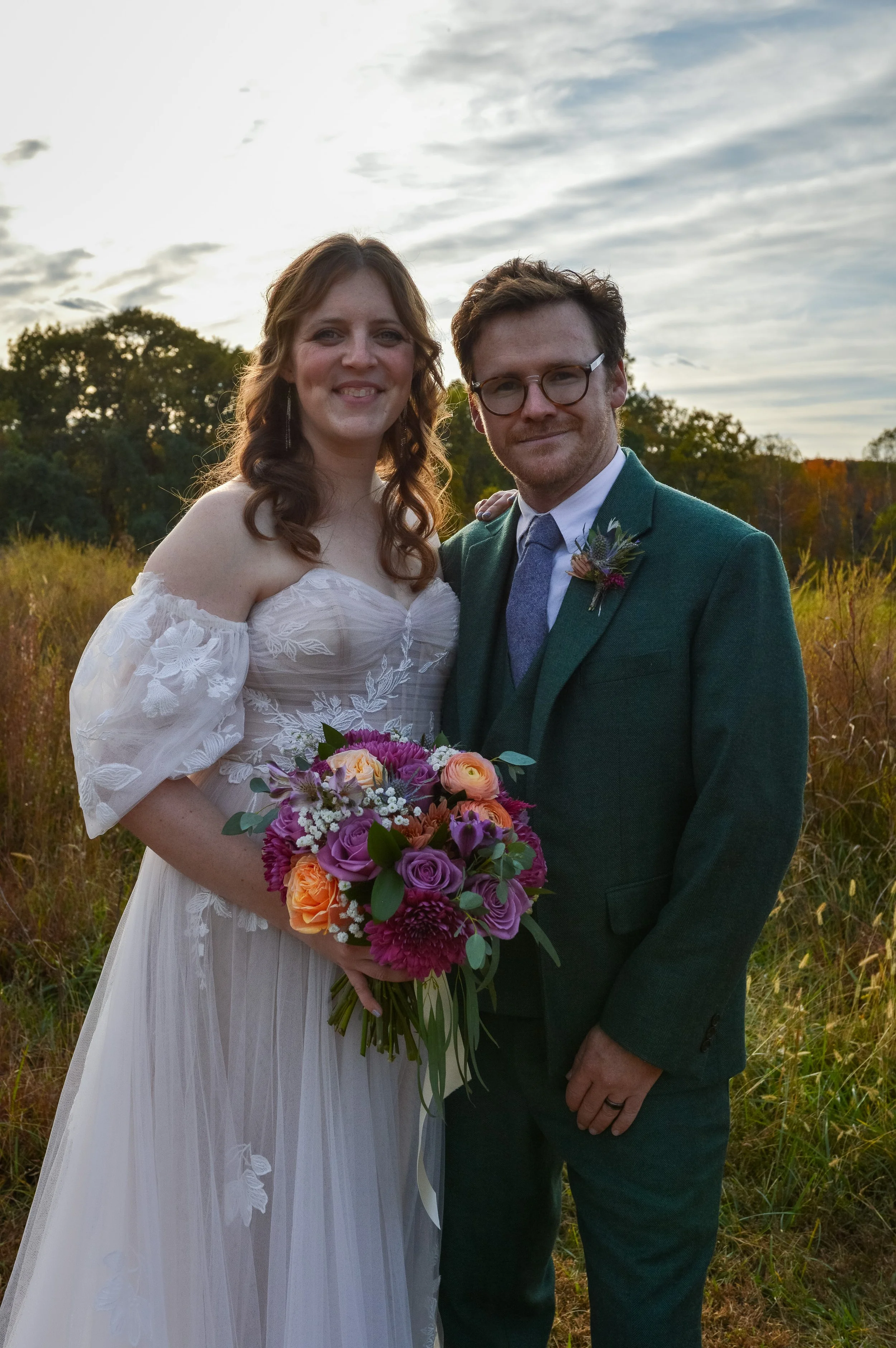 A bride and groom standing outdoors in a field with trees and cloudy sky in the background, smiling at the camera. The bride is holding a colorful bouquet of flowers, and the groom is dressed in a green suit with a boutonniere.