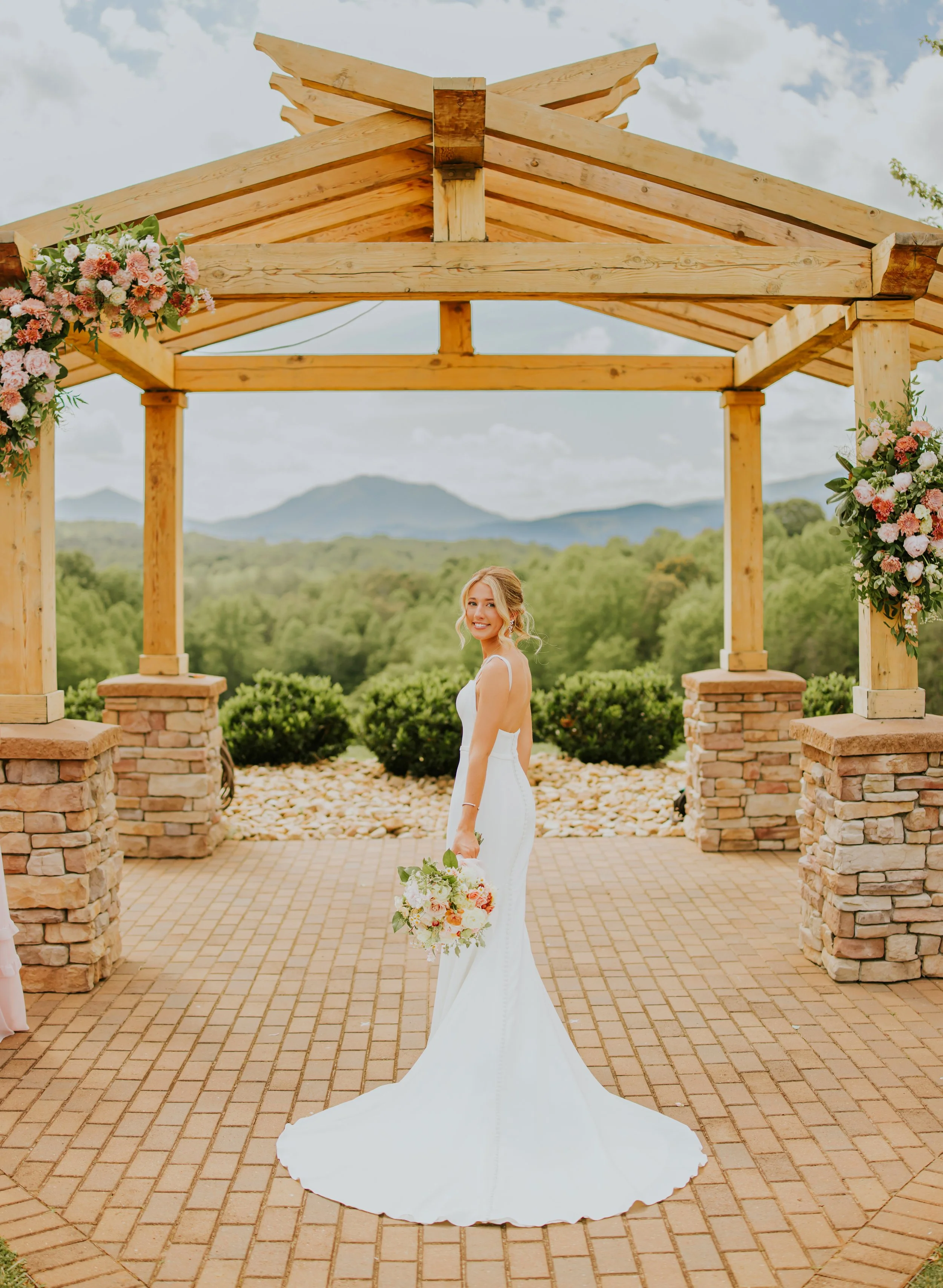A bride in a white wedding dress holding a bouquet of flowers, standing under a wooden arch decorated with pink and white flowers, with a scenic mountain view in the background.