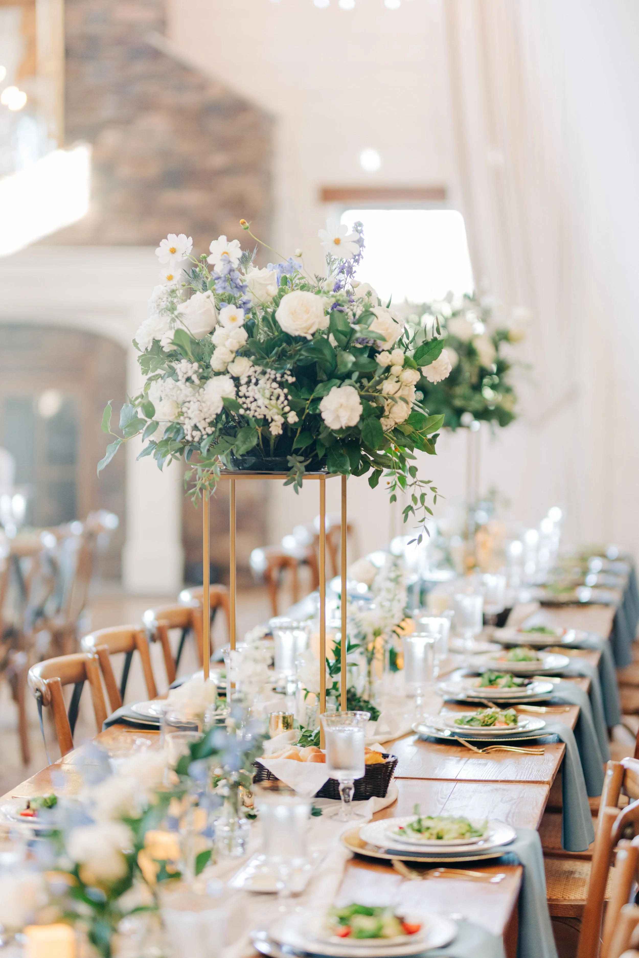 A long dining table set for a meal with plates, glasses, and cutlery, decorated with floral centerpieces of white and purple flowers on tall gold stands, in a bright room with curtains and wood accents.