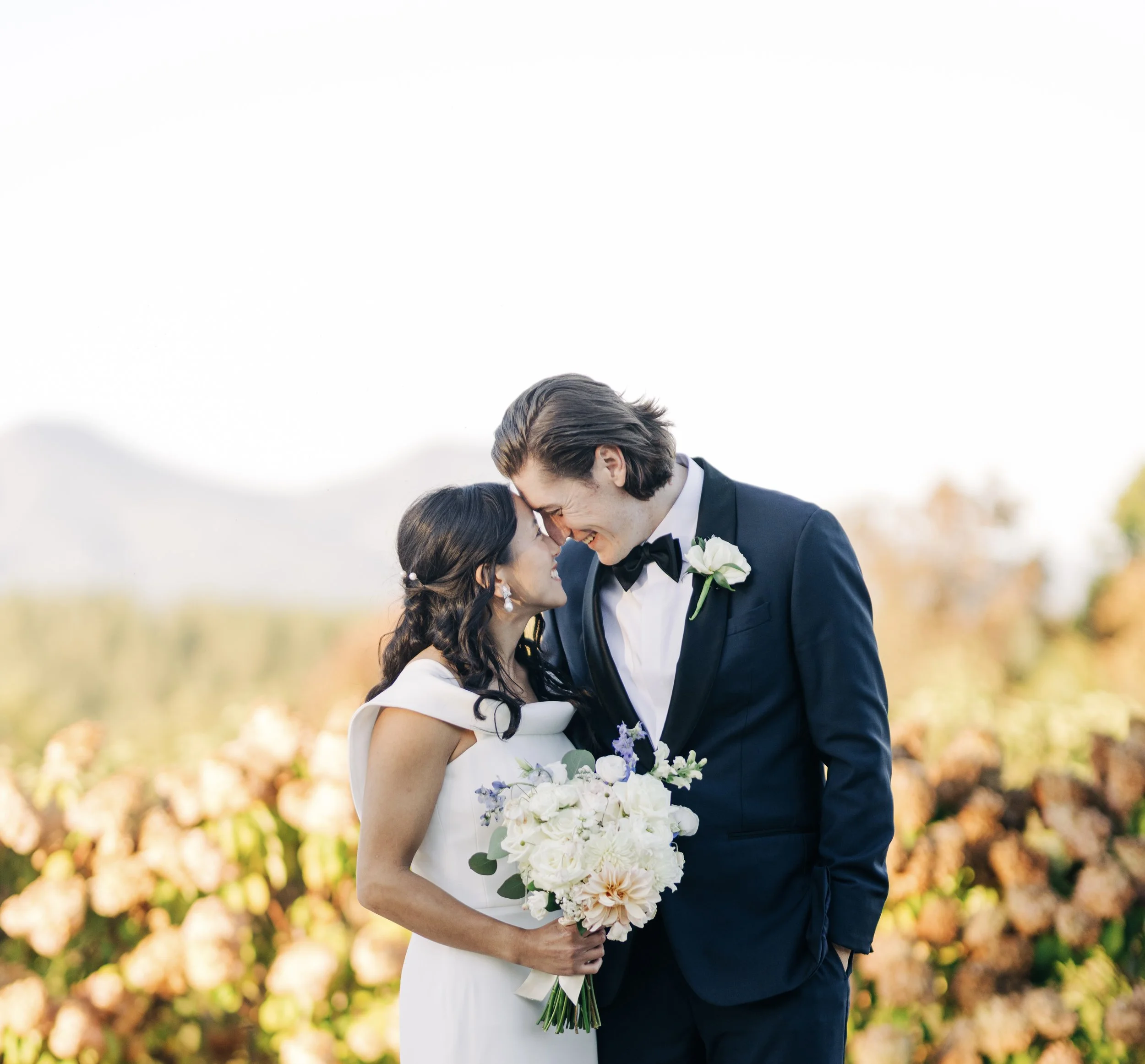 A bride and groom sharing a kiss outdoors during their wedding, with the bride holding a bouquet of white and purple flowers and the groom dressed in a tuxedo with a boutonniere.