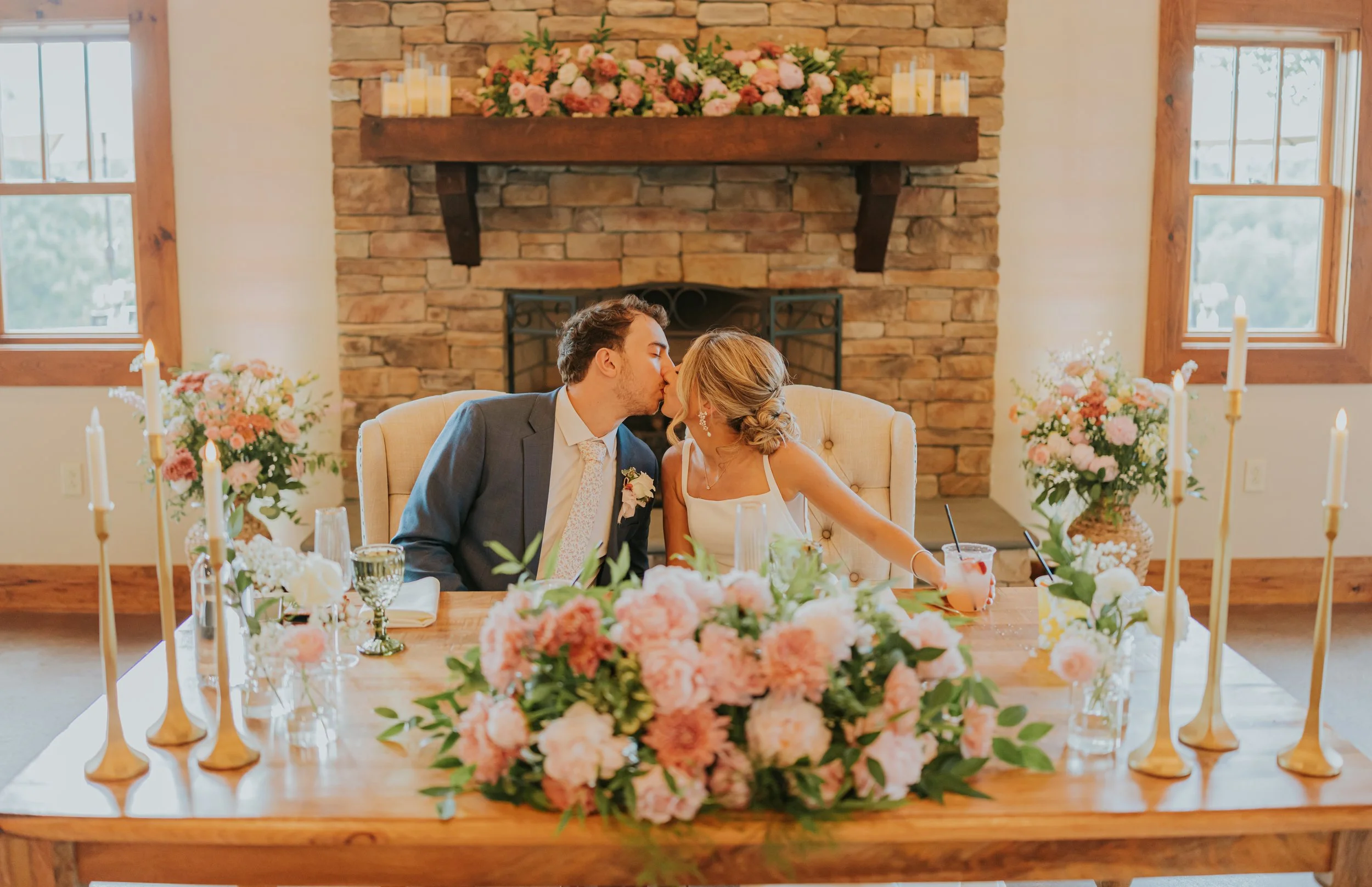 A couple is kissing at a wedding reception table decorated with pink and white flowers, candles, and glasses, set in a rustic room with brick fireplace and large windows.