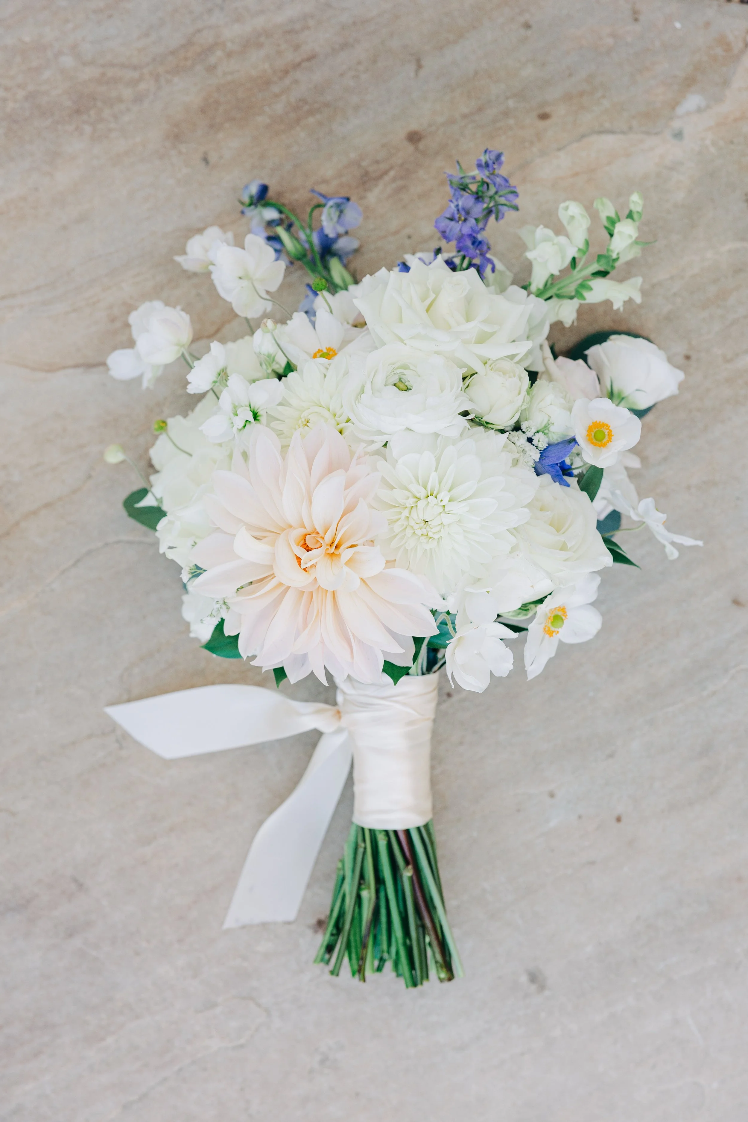 A bridal bouquet with white, cream, and purple flowers tied with a white satin ribbon and bow, resting on a beige textured surface.