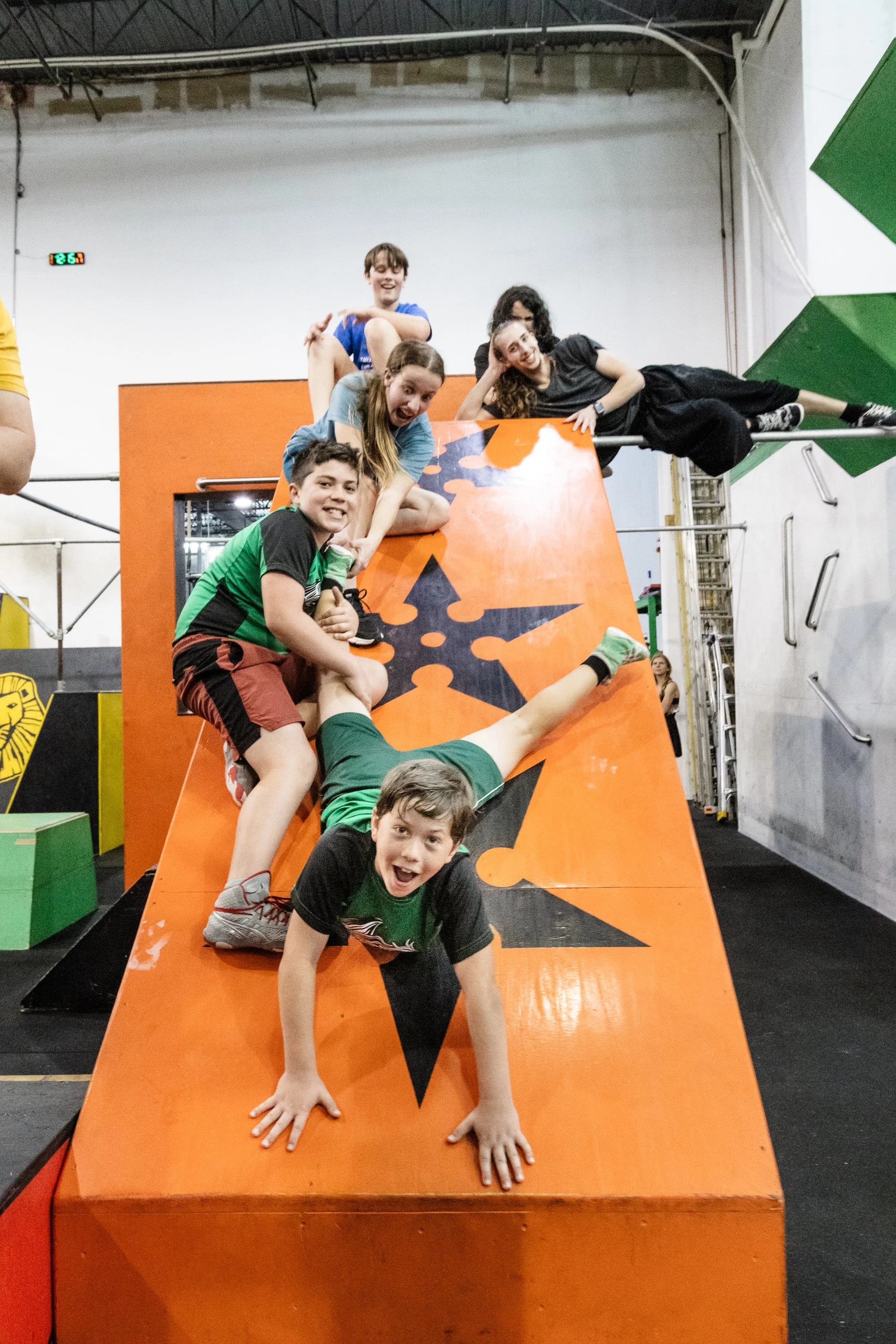 Indoor playground with a group of people, including a child in a colorful shirt posing with an instructor in a black tank top. Children playing on gym equipment and foam structures in the background.