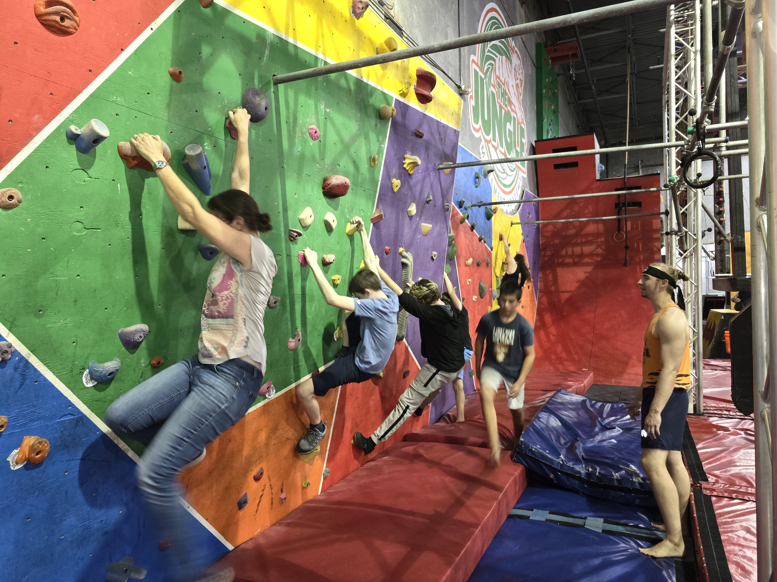 Child rock climbing in an indoor climbing gym, wearing a pink shirt and safety harness, using colorful climbing holds on the wall.