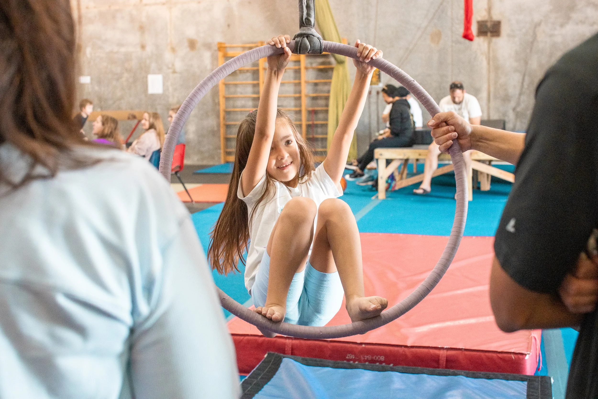 A group of people gathered in an indoor gym or training facility with obstacle courses. Some individuals are standing or sitting on various structures, including ramps and platforms. The setting has athletic equipment and appears to host a class, group activity, or event.