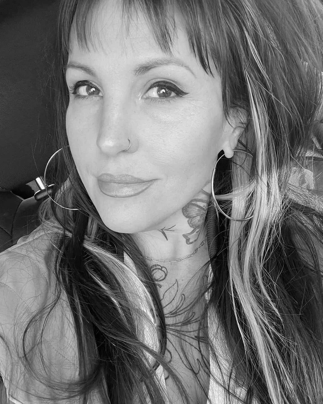 A Black and white portrait of a woman with large hoop earrings, a nose ring, tattoos on her neck and chest, and long wavy hair.